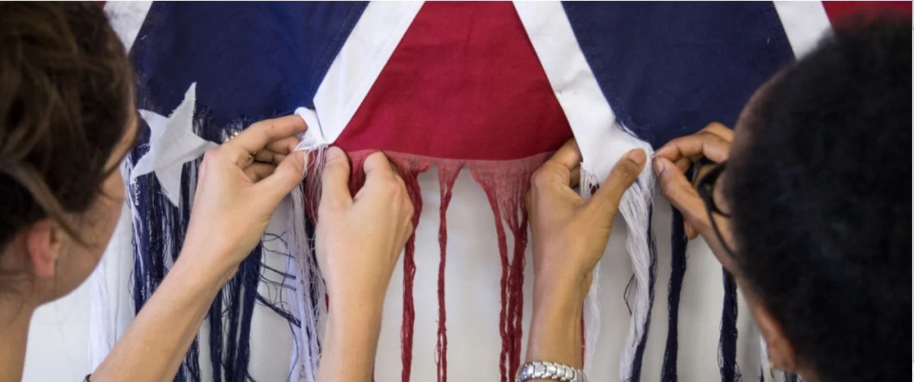 Sonya Clark and a visitor unpicking the flag. Unraveling , Sonya Clark, 2015, Cotton Confederate Flag. Photo: Courtesy The Artist. Photo Taylor Dabney