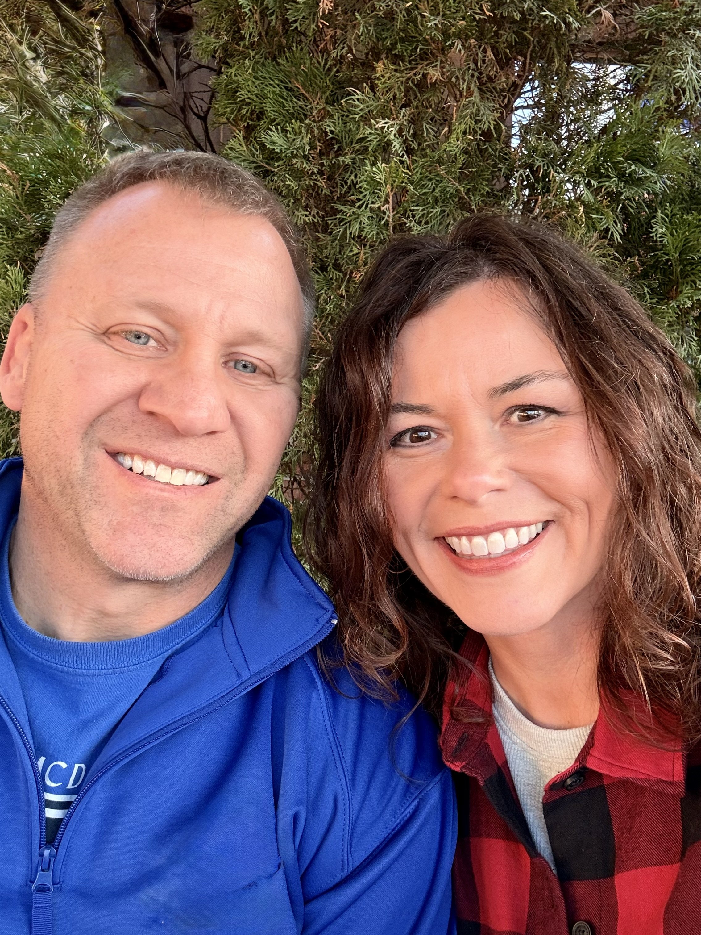A close-up photo of a smiling man and woman outdoors, standing near a green leafy shrub, with the man on the left and the woman on the right.
