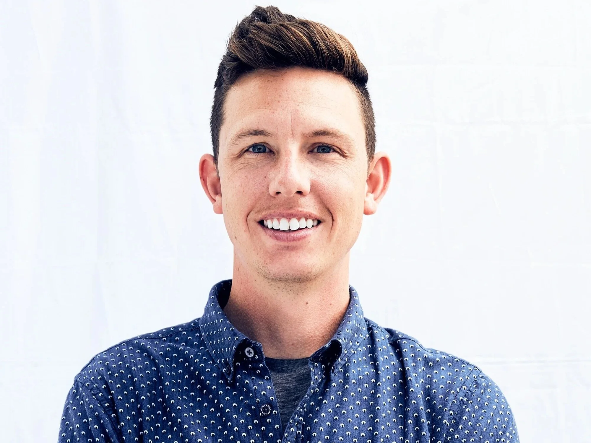 A young man with short brown hair and blue eyes smiling, wearing a blue patterned button-up shirt, standing against a white background.