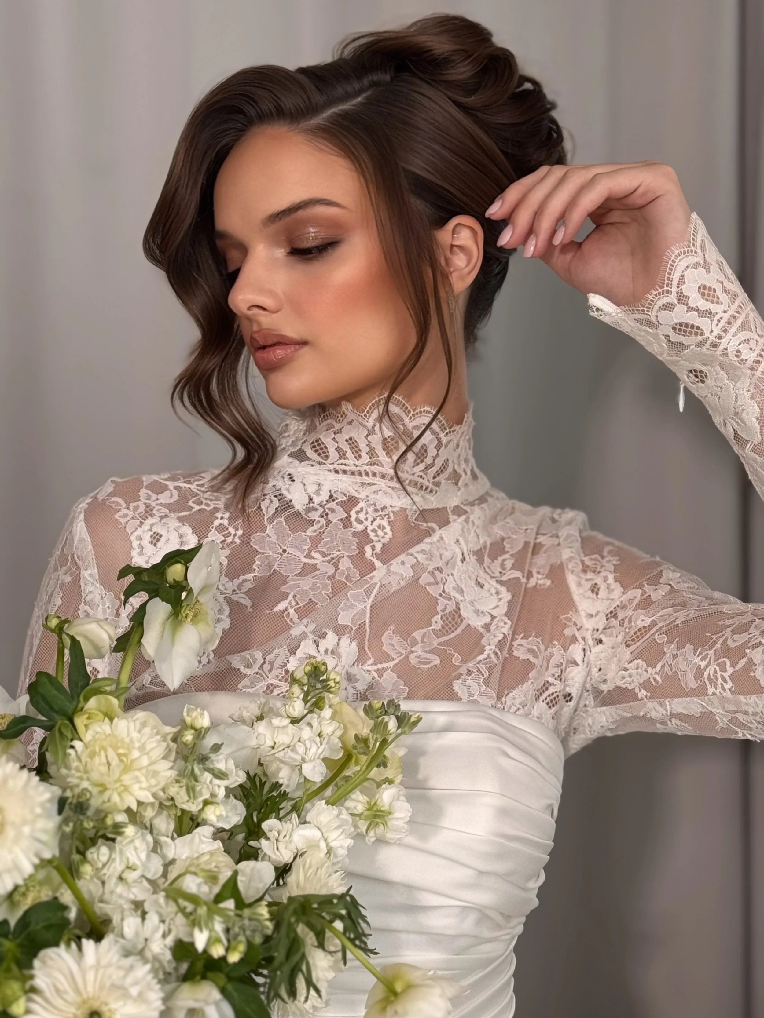 A woman with styled brown hair and makeup, wearing a white wedding dress with lace details, holds a bouquet of white flowers and looks downward with her eyes closed.