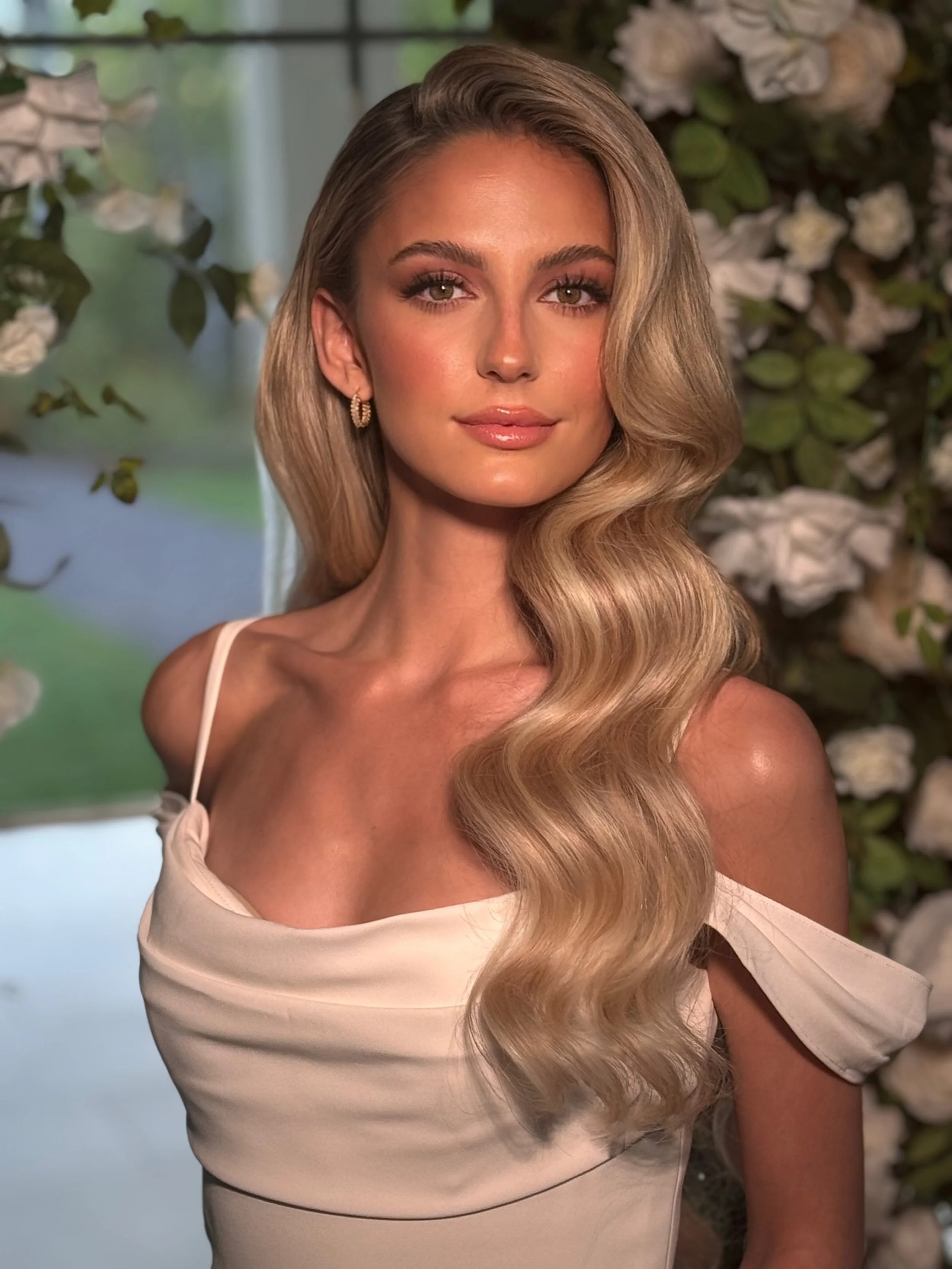 A woman with long, wavy blonde hair wearing a cream-colored dress with a spaghetti strap and earrings, standing in front of a floral backdrop.
