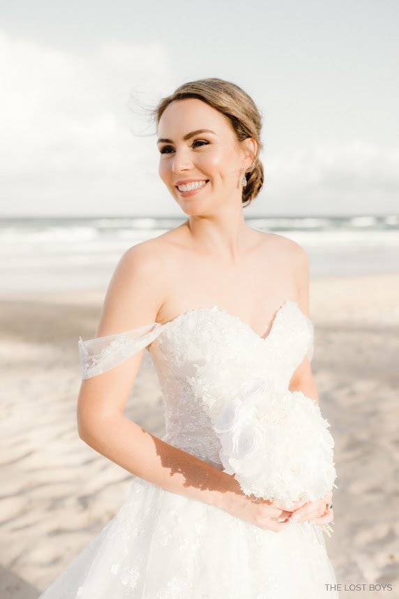 A happy bride on a Sunshine Coast beach with a floral bouquet, featuring natural glowy makeup designed for bright, outdoor coastal light.
