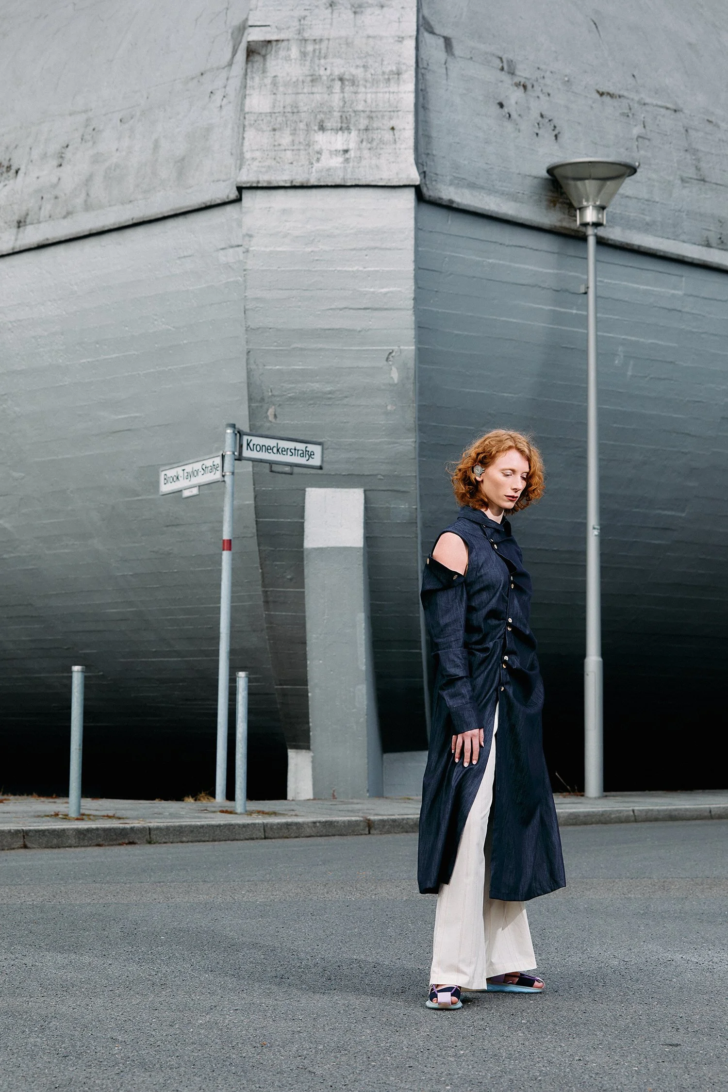 A young woman with red, curly hair stands on a street corner in Berlin, Germany, in front of a large, modern, gray building façade, wearing a dark, longer jacket with open shoulders, white pants and open-toed shoes.