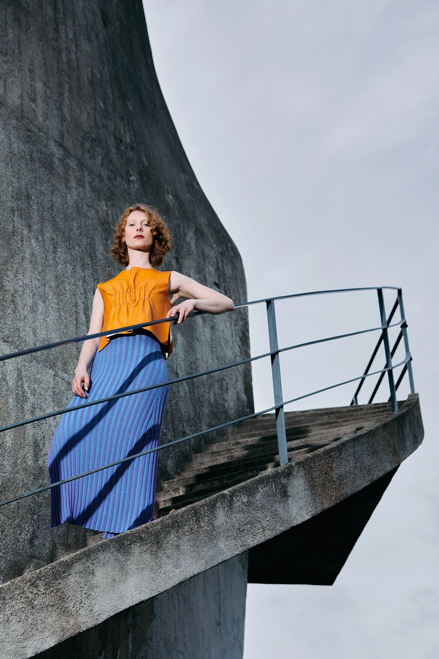 A woman stands on a balcony in front of a large concrete wall, wearing an orange top and a long blue skirt with stripes, and looks into the camera.