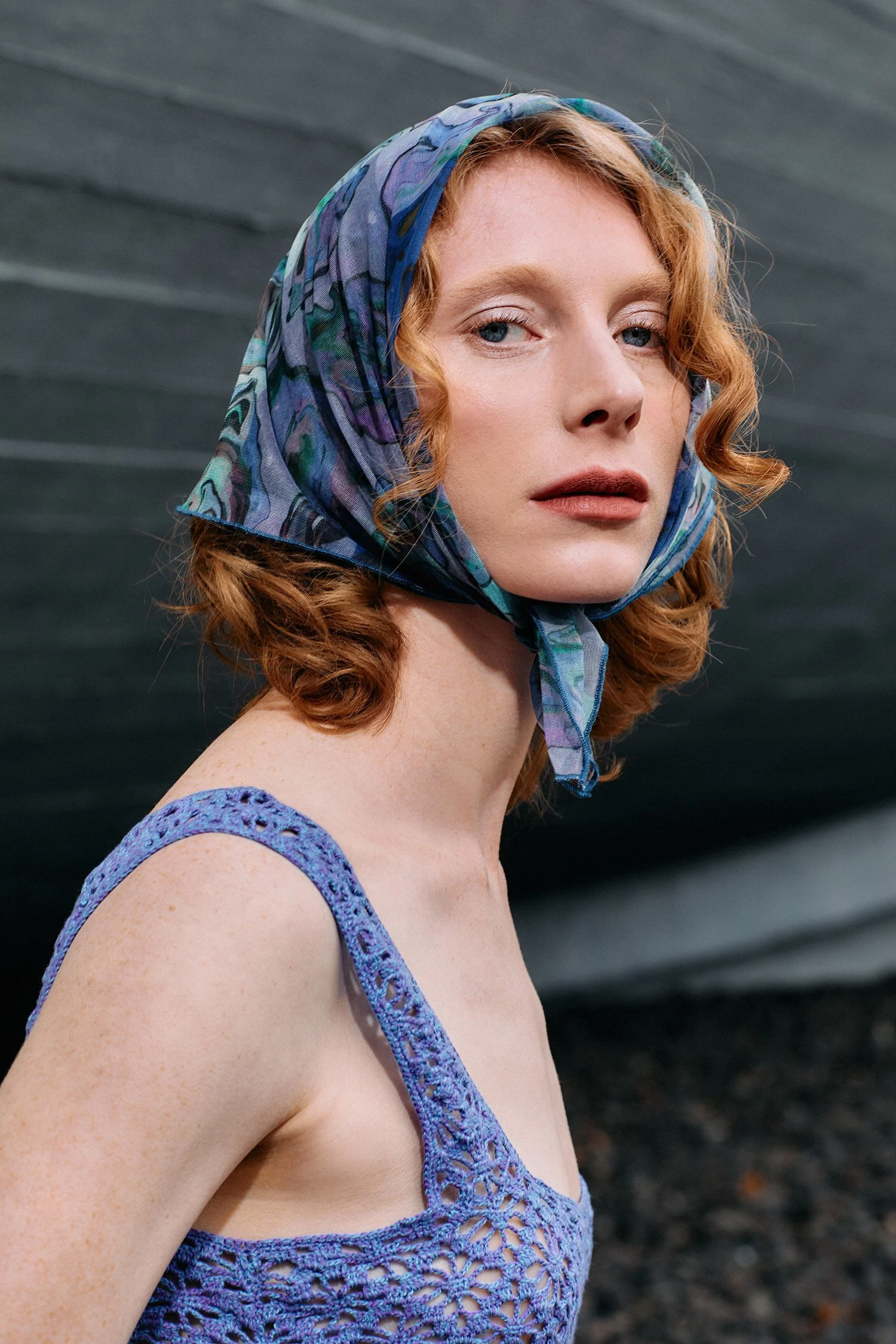 Young woman with red, curly hair wearing a colorful scarf on her head and a lavender crochet top, looking to the left, a dark wall in the background.