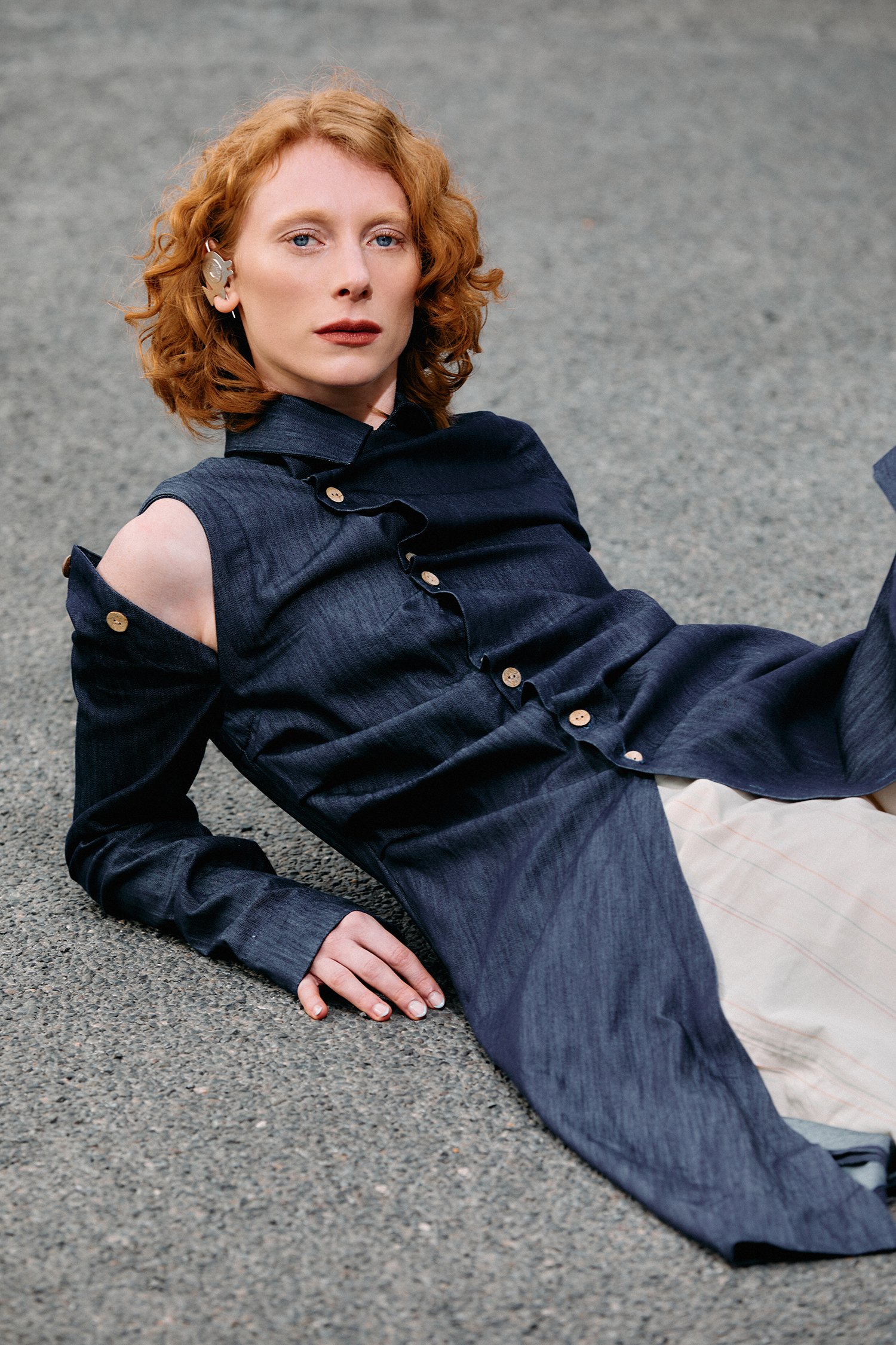 A red-haired woman is lying on a street surface, wearing a dark blue open-shouldered shirt jacket and beige pants, looking directly into the camera.