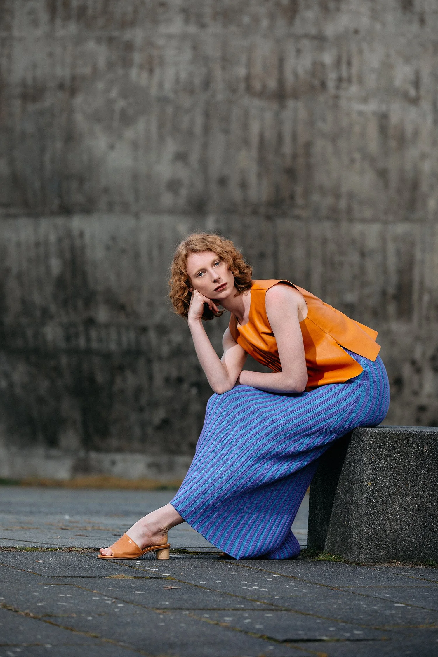 A woman with curly red hair, wearing a colorful outfit, sitting on a stone bench in an urban setting with a large concrete wall in the background.
