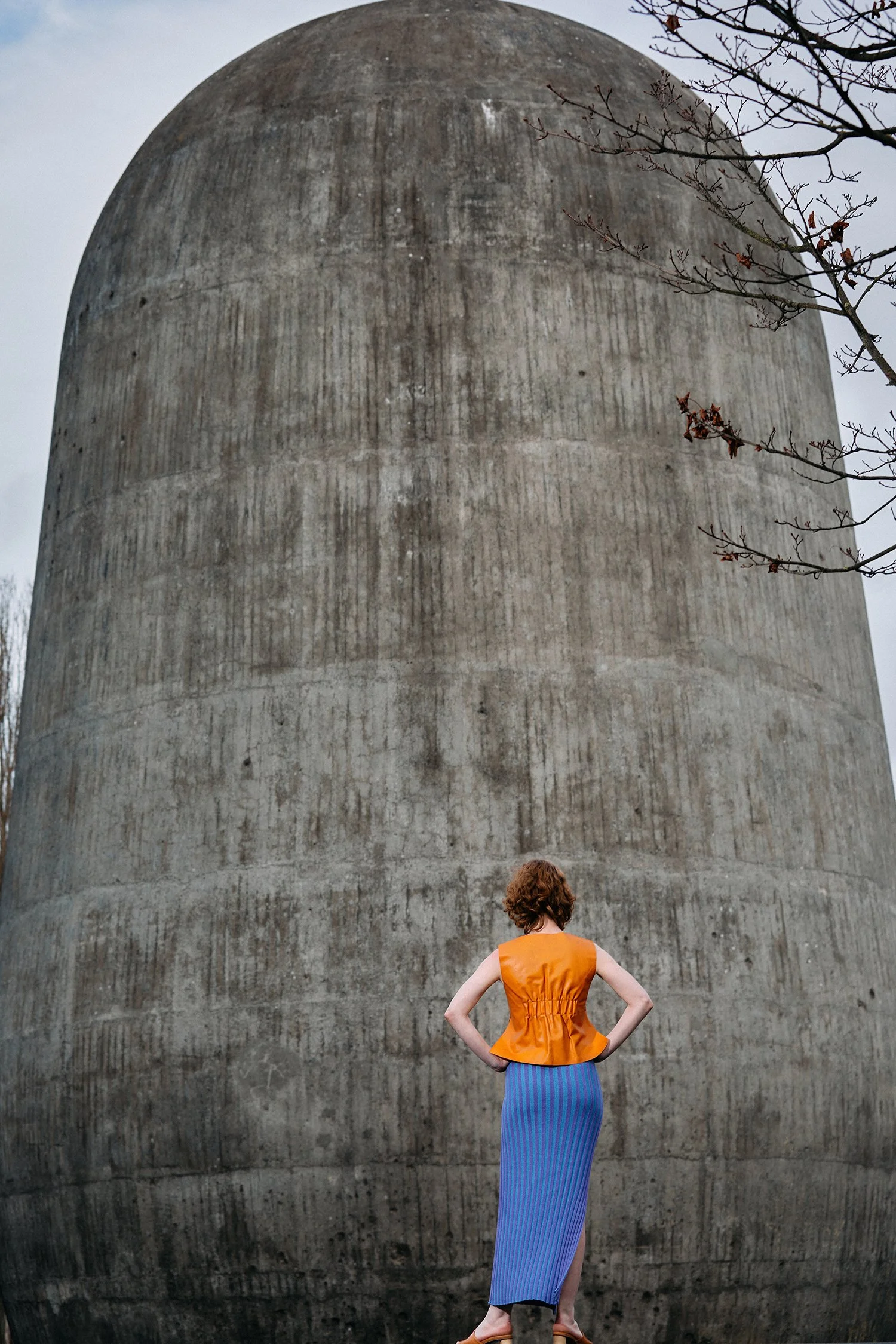 A woman stands in front of a large, round concrete wall outside, wearing an orange top and a blue, striped skirt, and looks up at the wall.