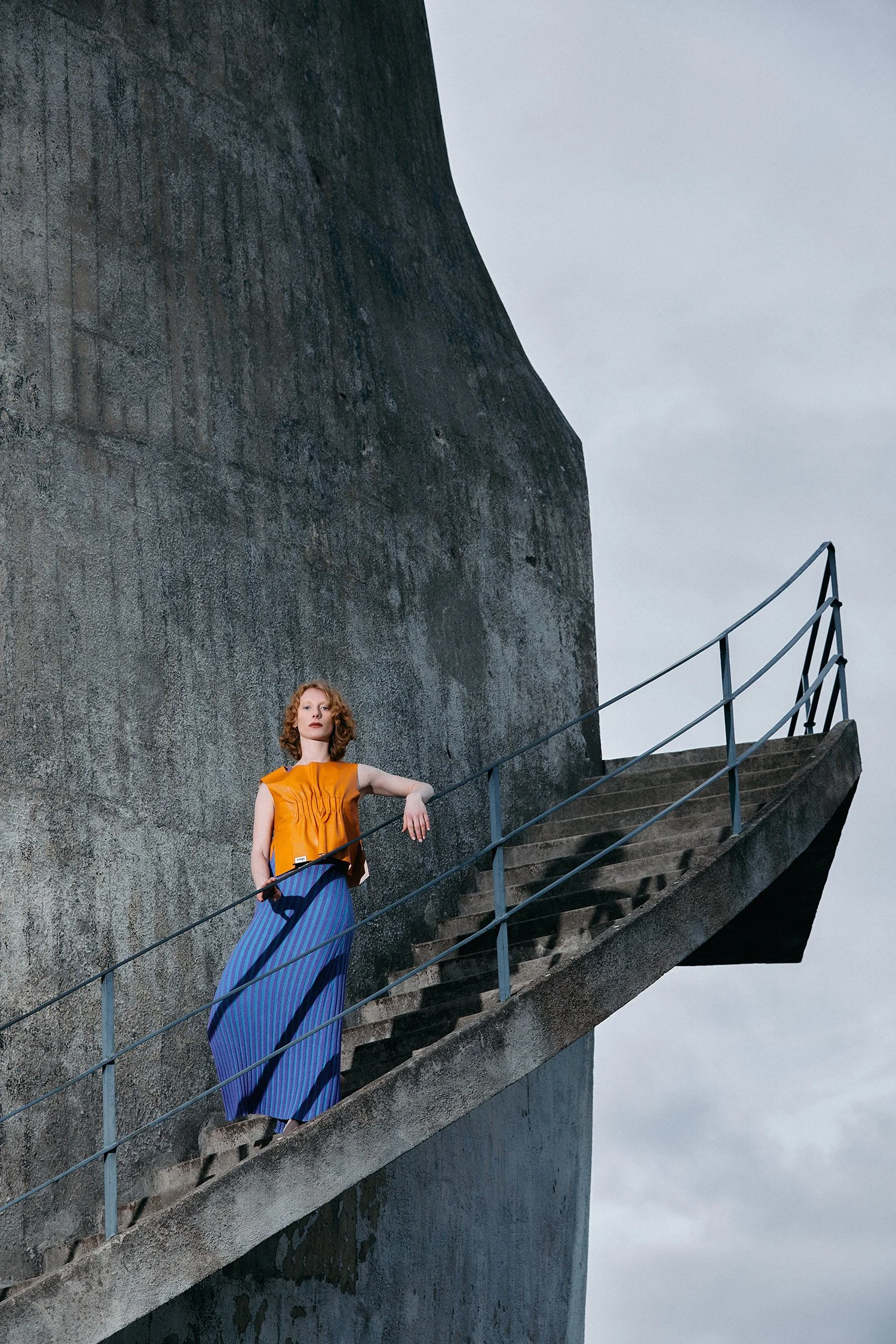 Young woman in a colorful dress stands on a staircase against a large concrete wall.