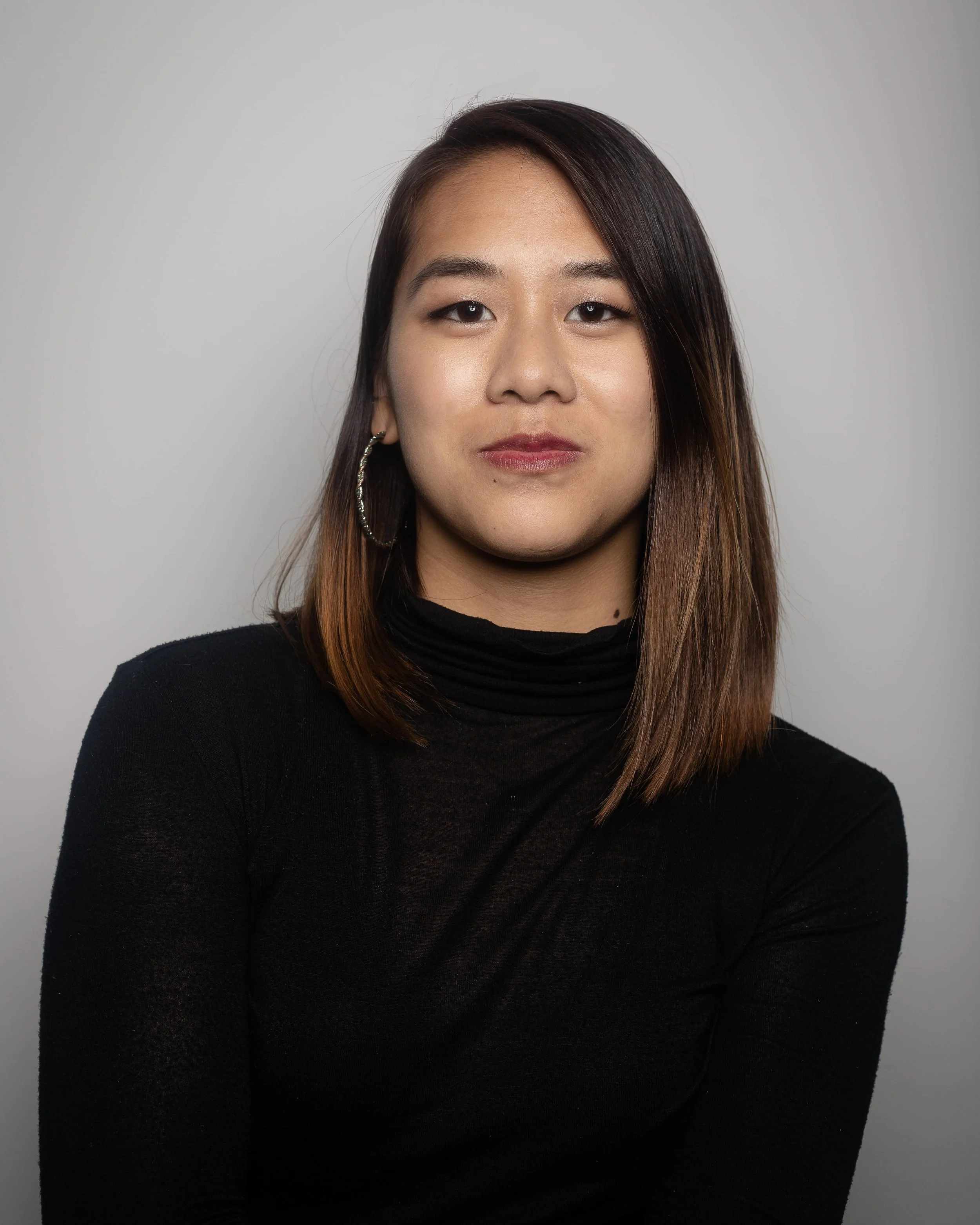 A young woman with long dark hair, wearing a black turtleneck, posing against a plain white background.