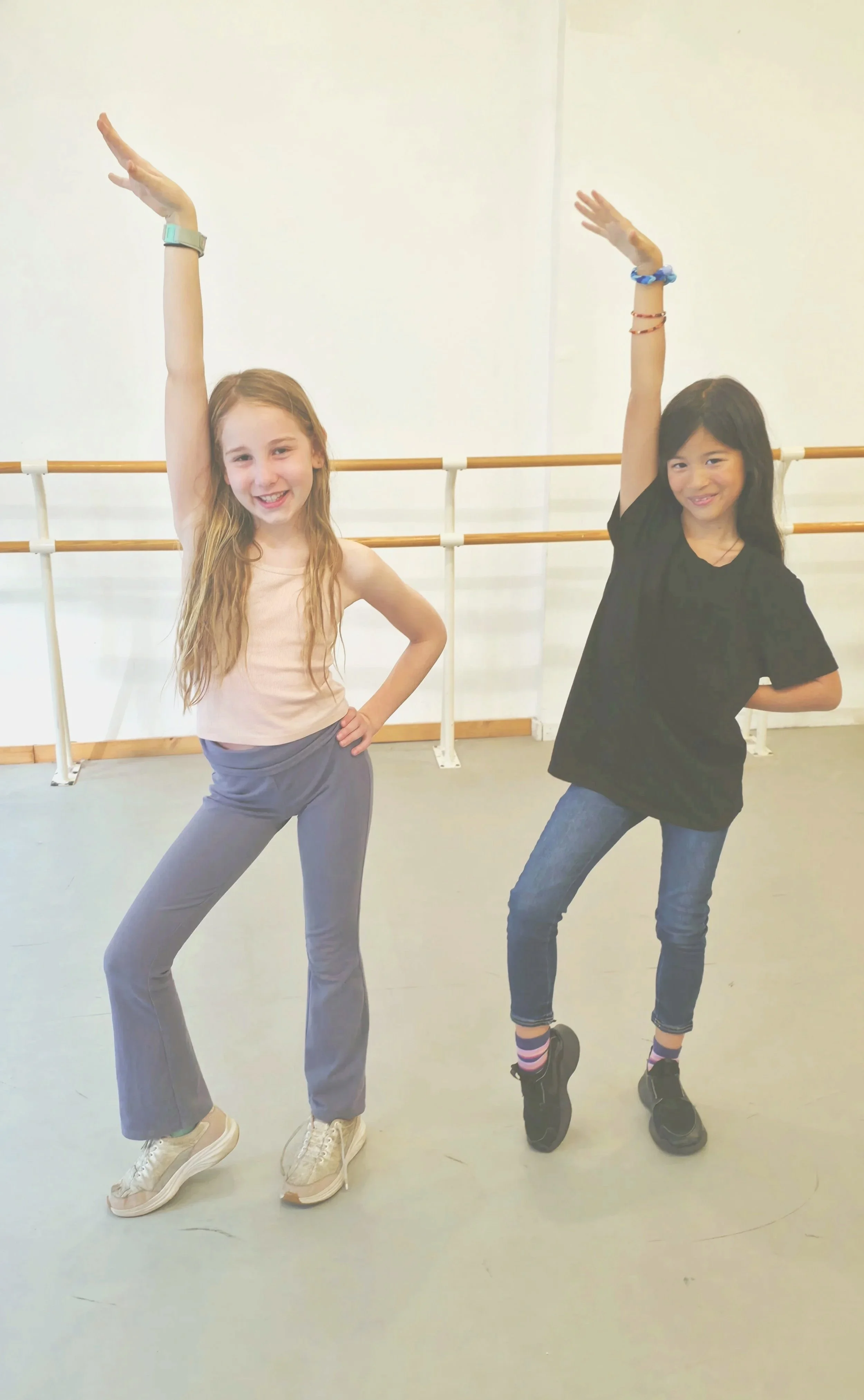 Two young girls pose in a dance studio with ballet barres, smiling and raising their arms in a dance pose.