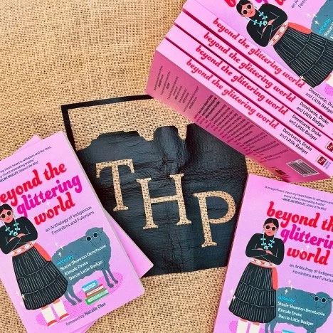 An image of the Torrey House Press logo, black stenciling on brown fabric, surrounded by pink paperbacks. The cover image of Beyond the Glittering World is a young Indigenous woman in a black blouse, turquoise concha, pink belt, & black skirt w mocs.