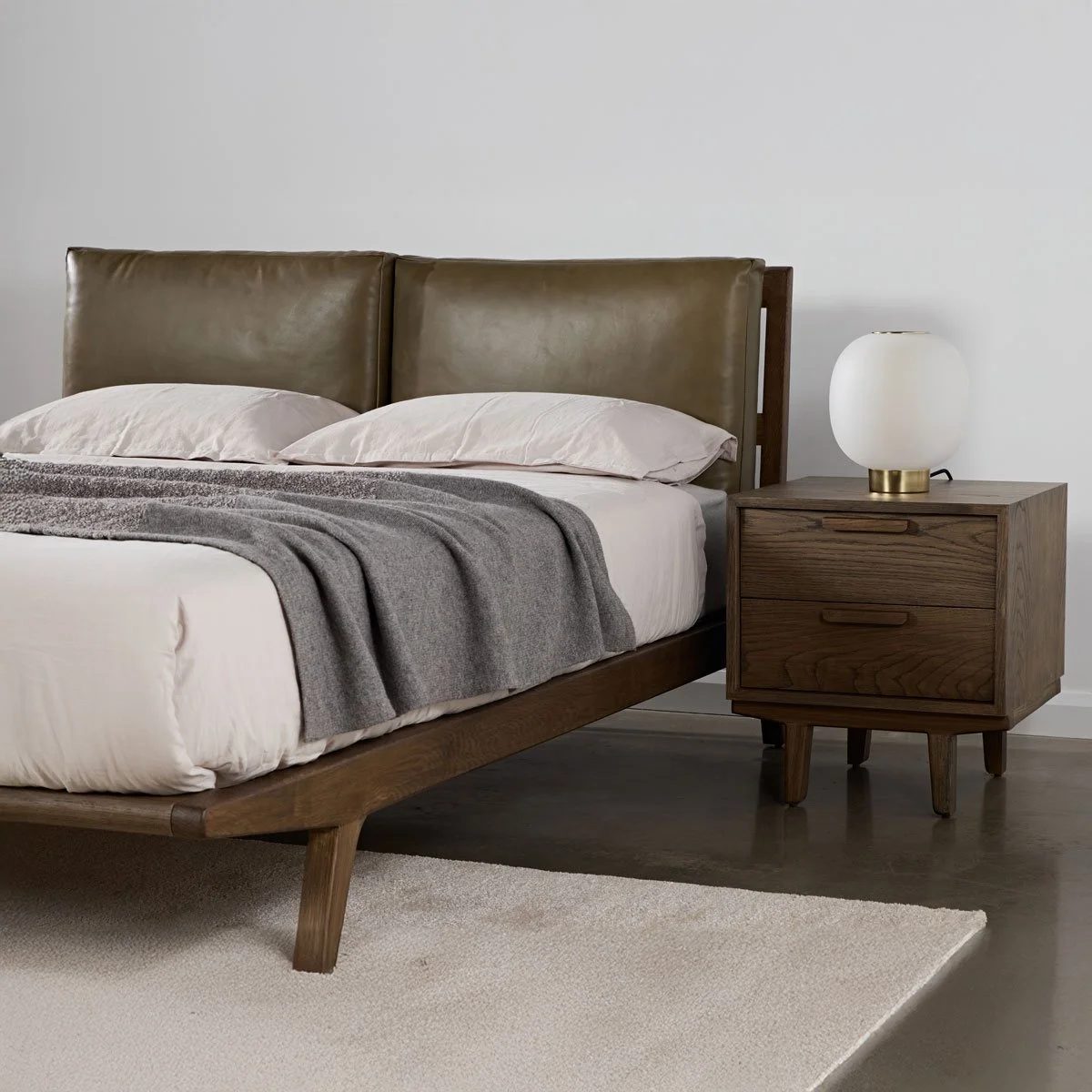 A bedroom with a wooden bed frame with a tan leather headboard, white pillows, a grey blanket, and a bedside wooden nightstand with a white lamp.