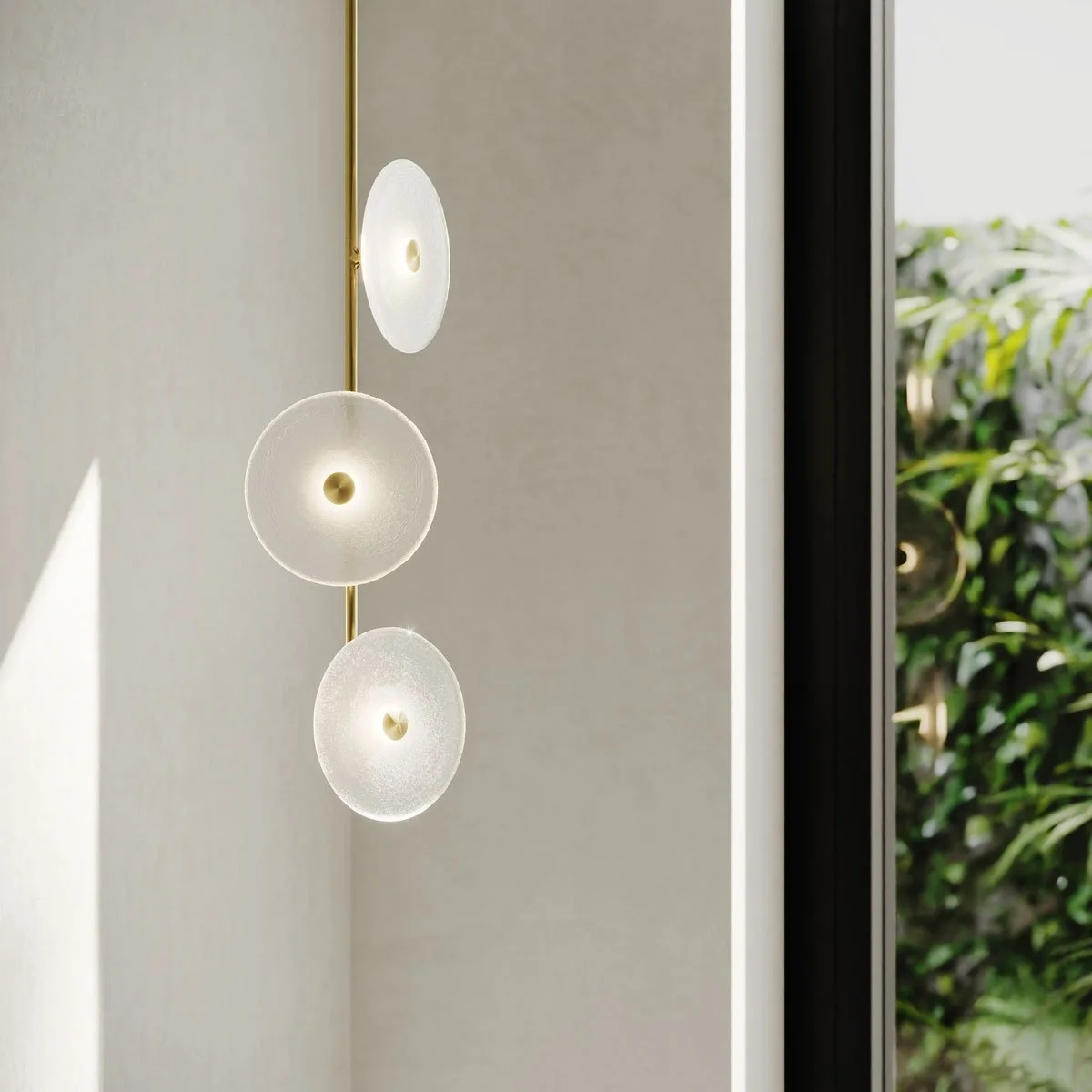 Modern wall-mounted light fixture with three translucent disc-shaped lights in front of a beige wall and near a window with greenery outside.