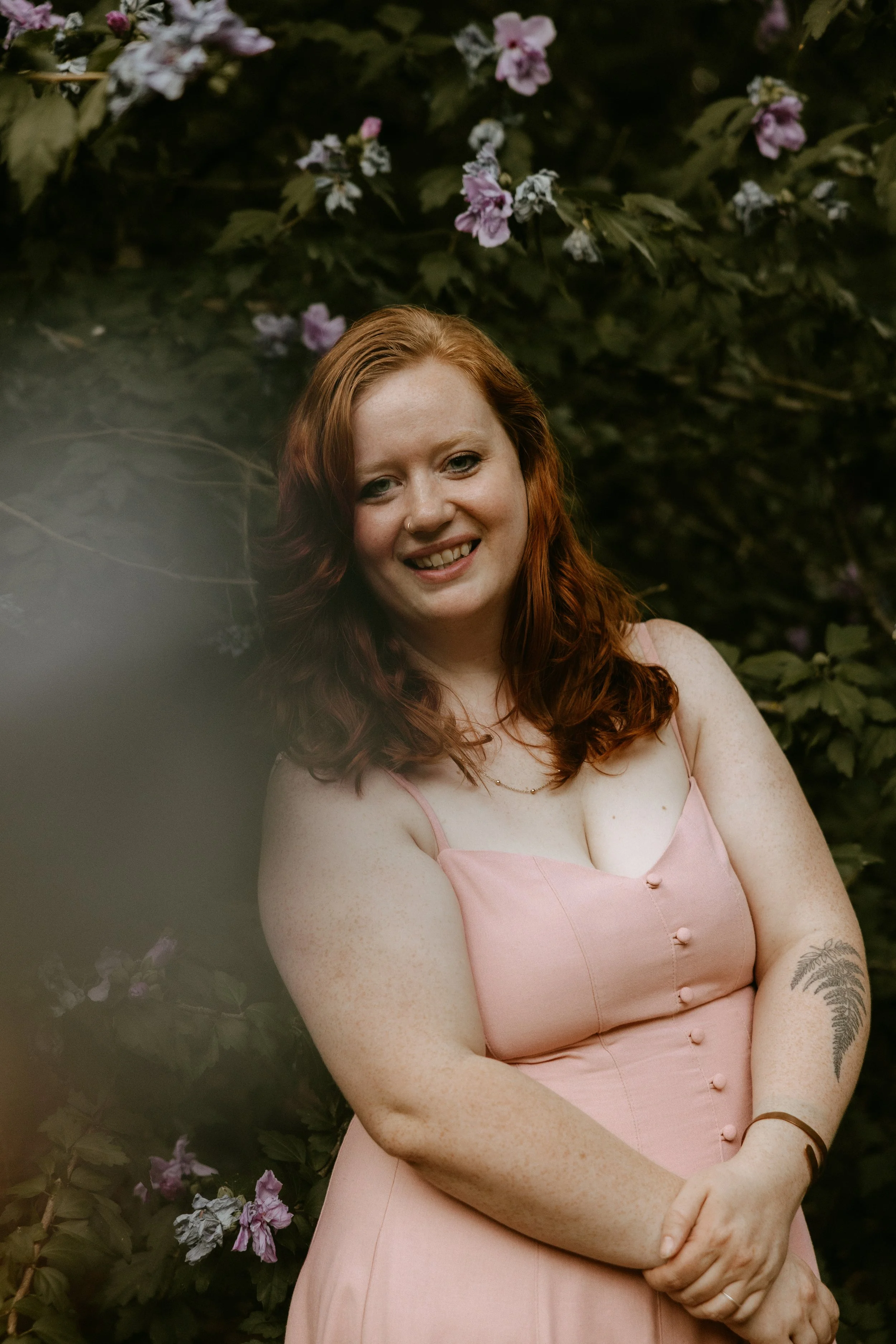 Photo of a woman with clasped hands smiling at the camera in front of a leafy bush with purple flowers.