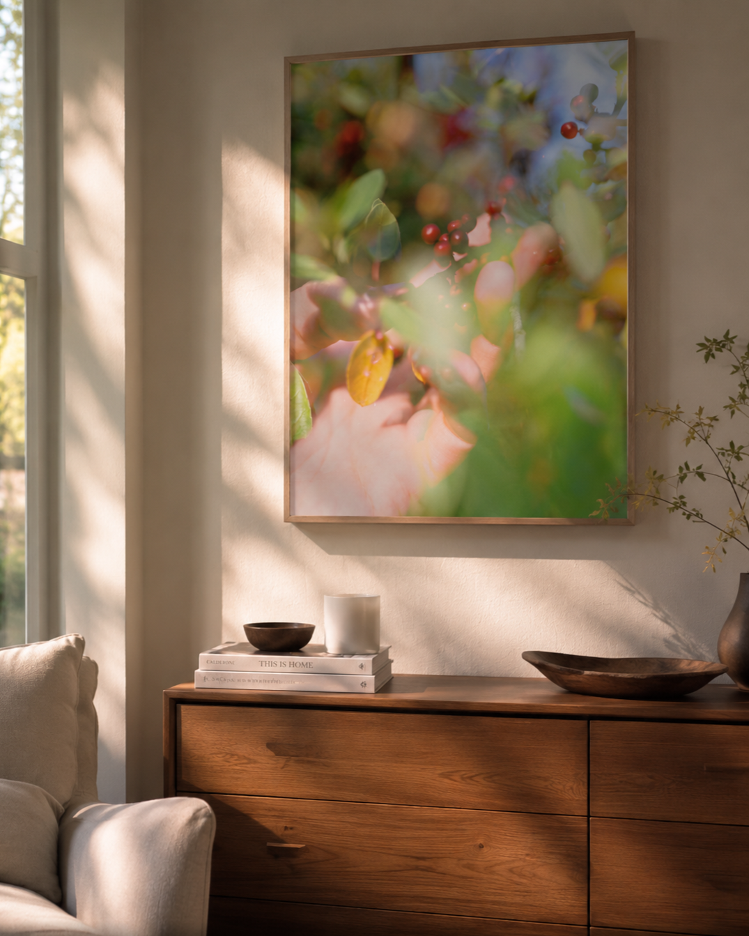 Interior of a living room decor with a large framed botanical print on the wall, a wooden sideboard with books and decorative items, sunlight streaming through a window on the left.