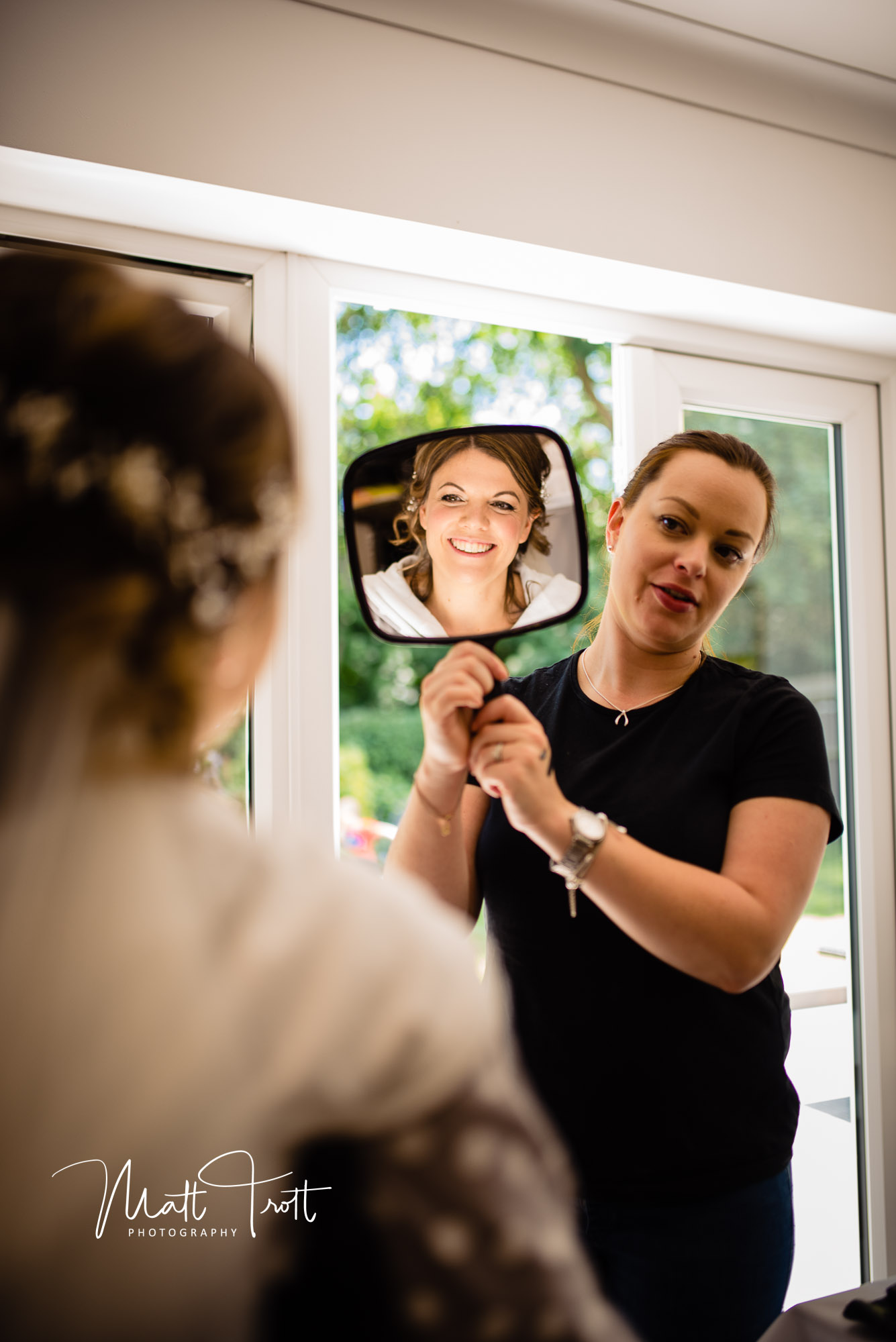 MUA holds up mirror for the sitting bride to see herself