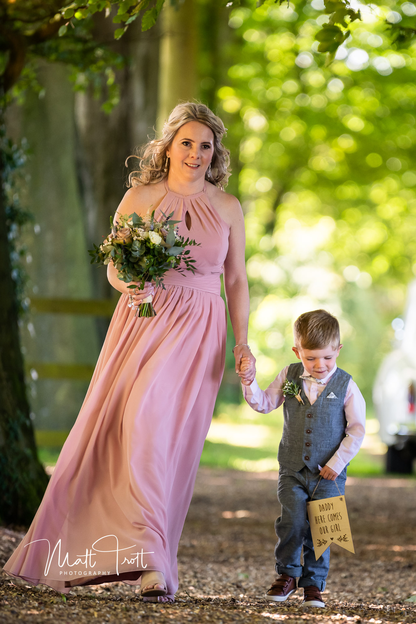 Head bridesmaid and young son of the bride walking down the aisle holding a sign saying here comes mummy
