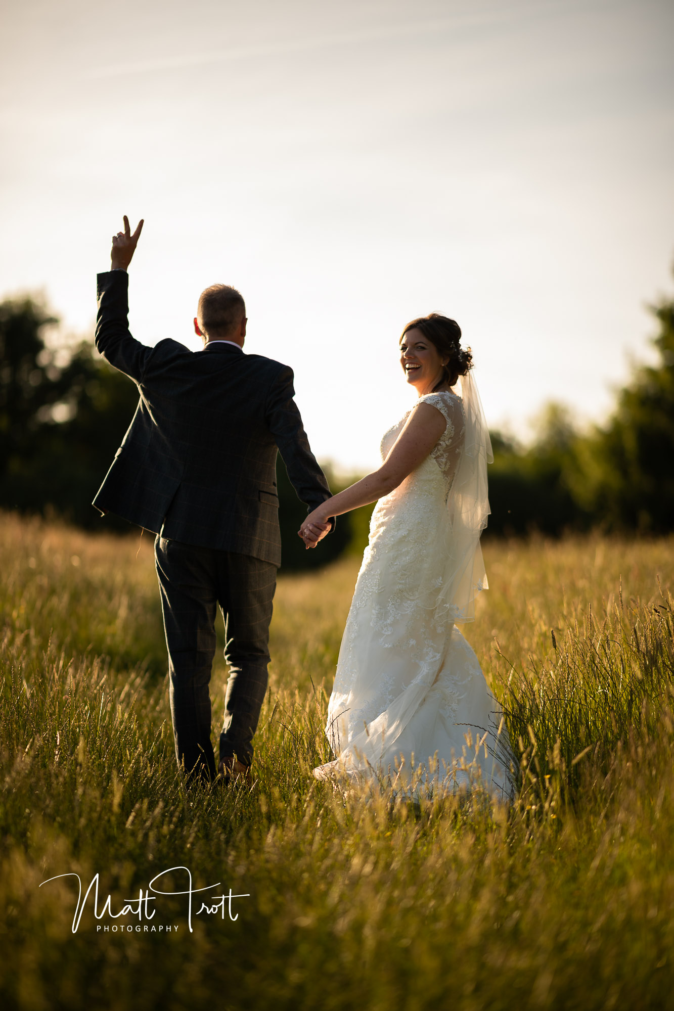 Groom gives the birdie whilst walking with wife through a sunset and long grass at crown lodge kent