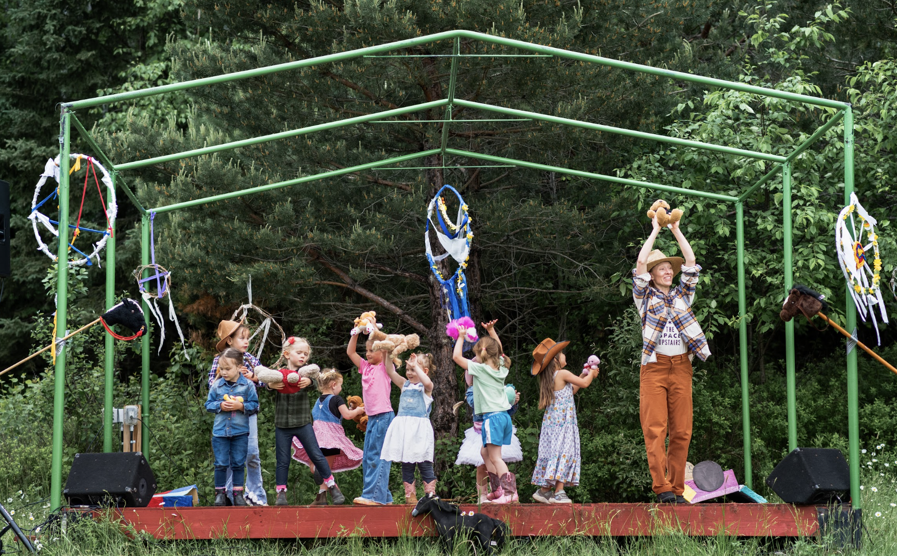 A group of youth artists and their teacher are standing on stage, holding teddy bears. There is green forest around them on the outdoor stage.