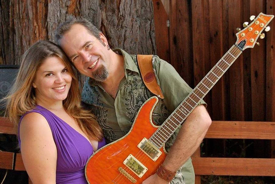 A smiling man with a guitar standing next to a smiling woman, outdoors near a wooden fence.
