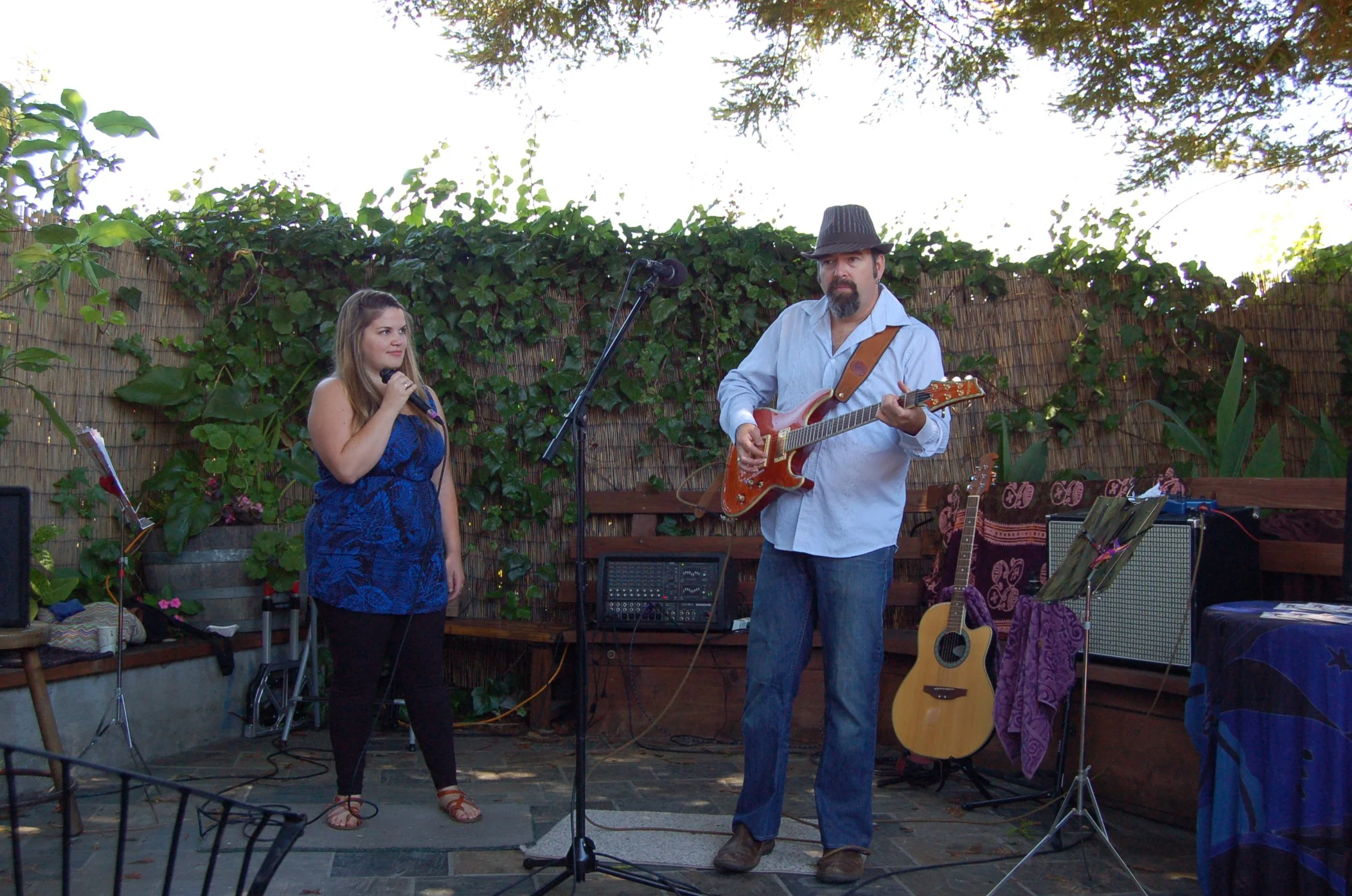 A man with a beard and mustache playing an electric guitar and a young woman holding a microphone on a small outdoor stage, with musical equipment and guitars around them, surrounded by green plants and a bamboo fence.