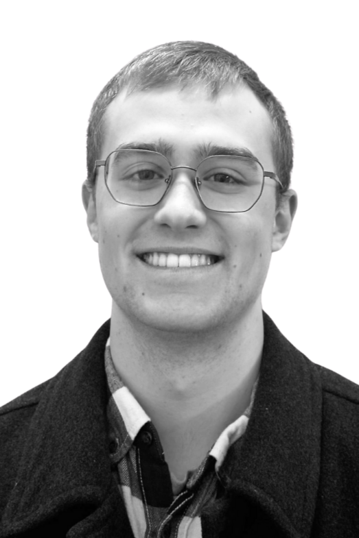 A young man with short dark hair, a beard and mustache, smiling, wearing a t-shirt and a necklace, against a plain wall.