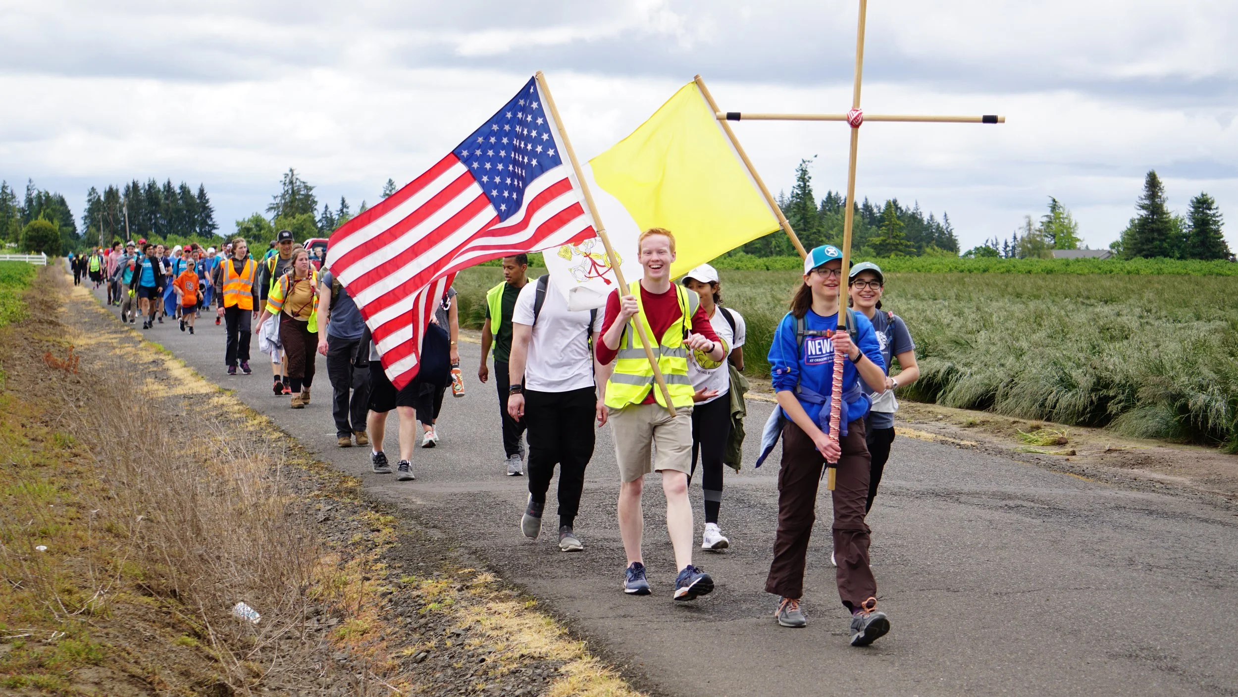 Group of people walking along a rural road, some carrying American flags, in an outdoor setting with fields and trees, under a cloudy sky.