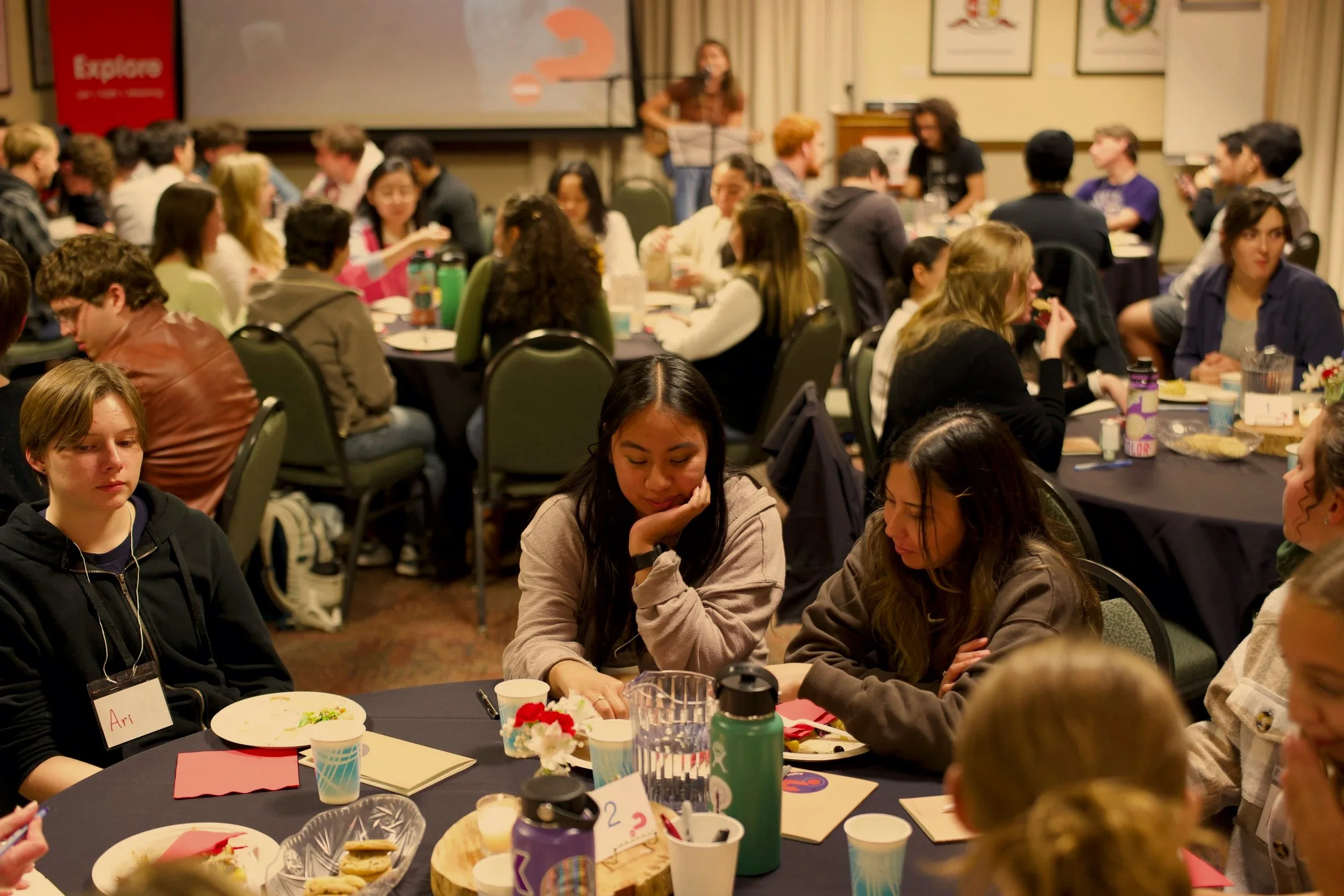 A large gathering of people seated at round tables in a conference room, with some engaged in conversation and others eating. In the background, a woman is standing at a podium and speaking.