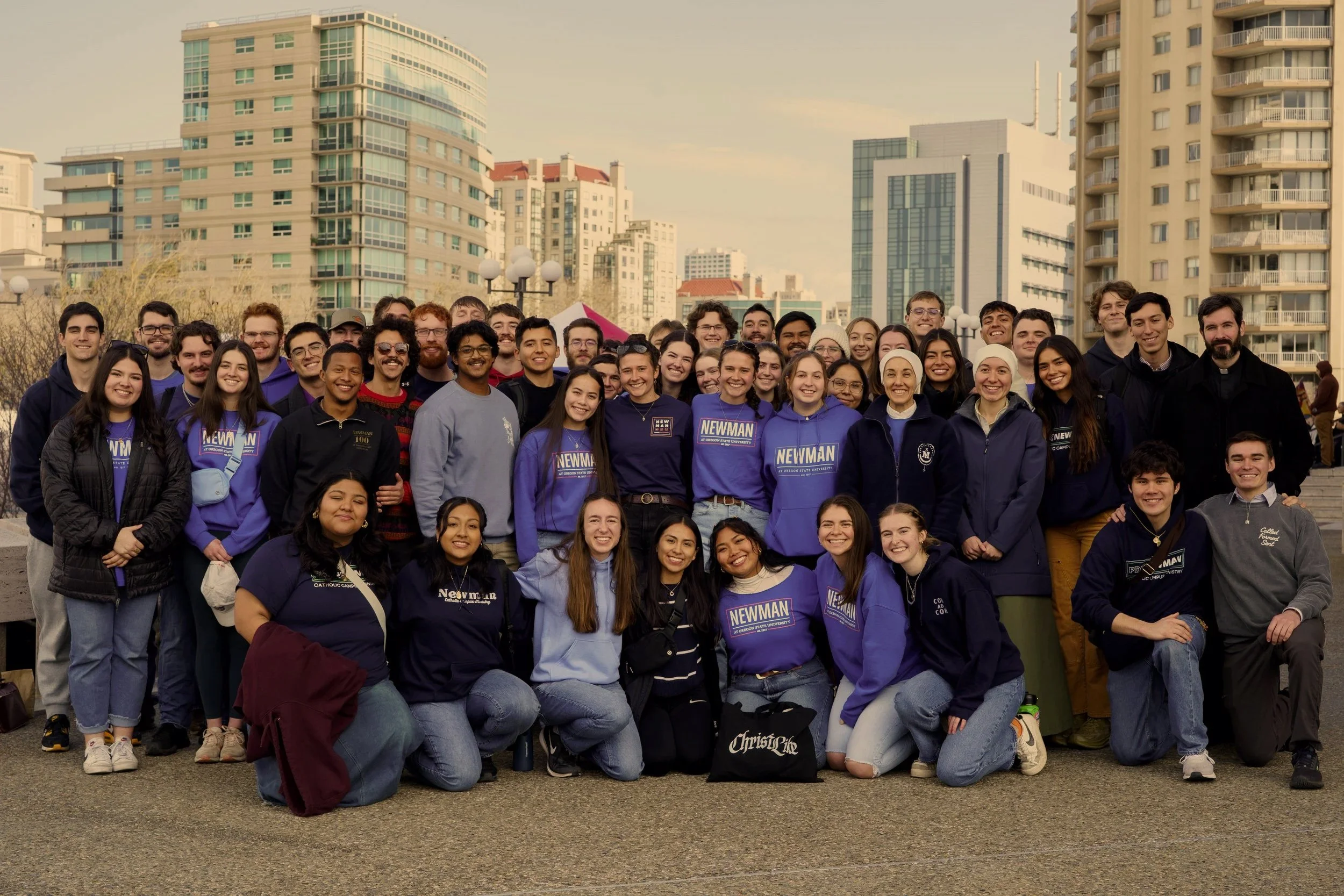 A large group of diverse young adults posing together outdoors in an urban setting. Some are wearing purple or blue shirts with 'NEWMAN' on them, indicating they are part of the same organization or event. The background features modern high-rise bui