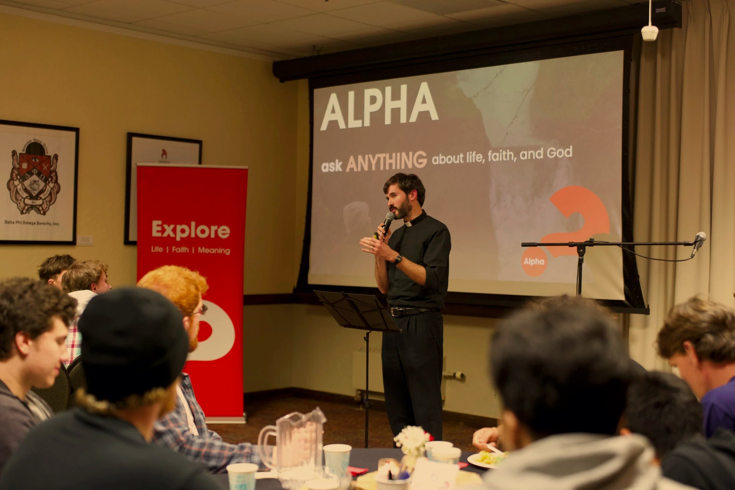 A priest speaking into a microphone during a presentation titled "ALPHA" about ask anything related to life, faith, and God. The audience is seated at tables with cups and pitchers, and a red banner with the words "Explore Life Faith Meaning" is visi