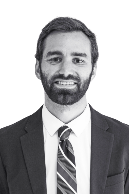 Black and white professional headshot of a man wearing a suit, tie, and white shirt, with short hair and a beard, against a plain background.