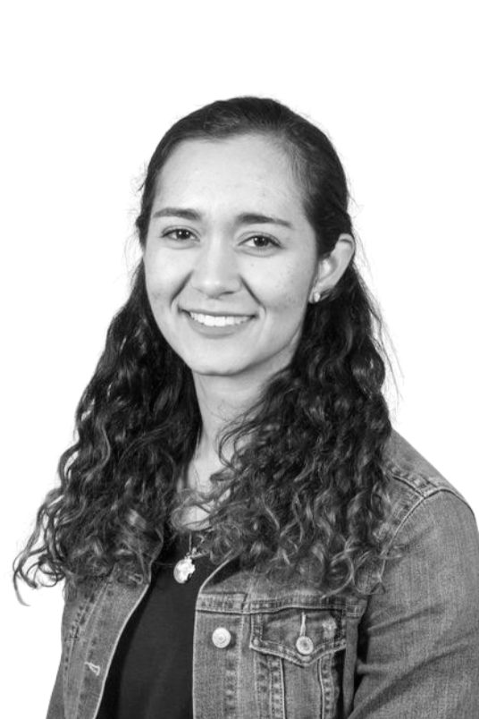 Black and white portrait of a young woman with shoulder-length wavy hair, smiling, wearing a polka-dotted top, standing against a plain background.