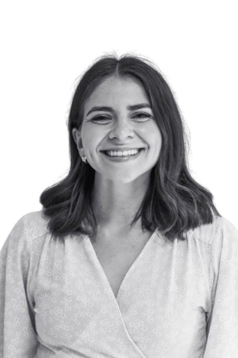 A woman with medium-length dark hair, smiling, wearing a light-colored blouse with a floral pattern, standing against a plain background.