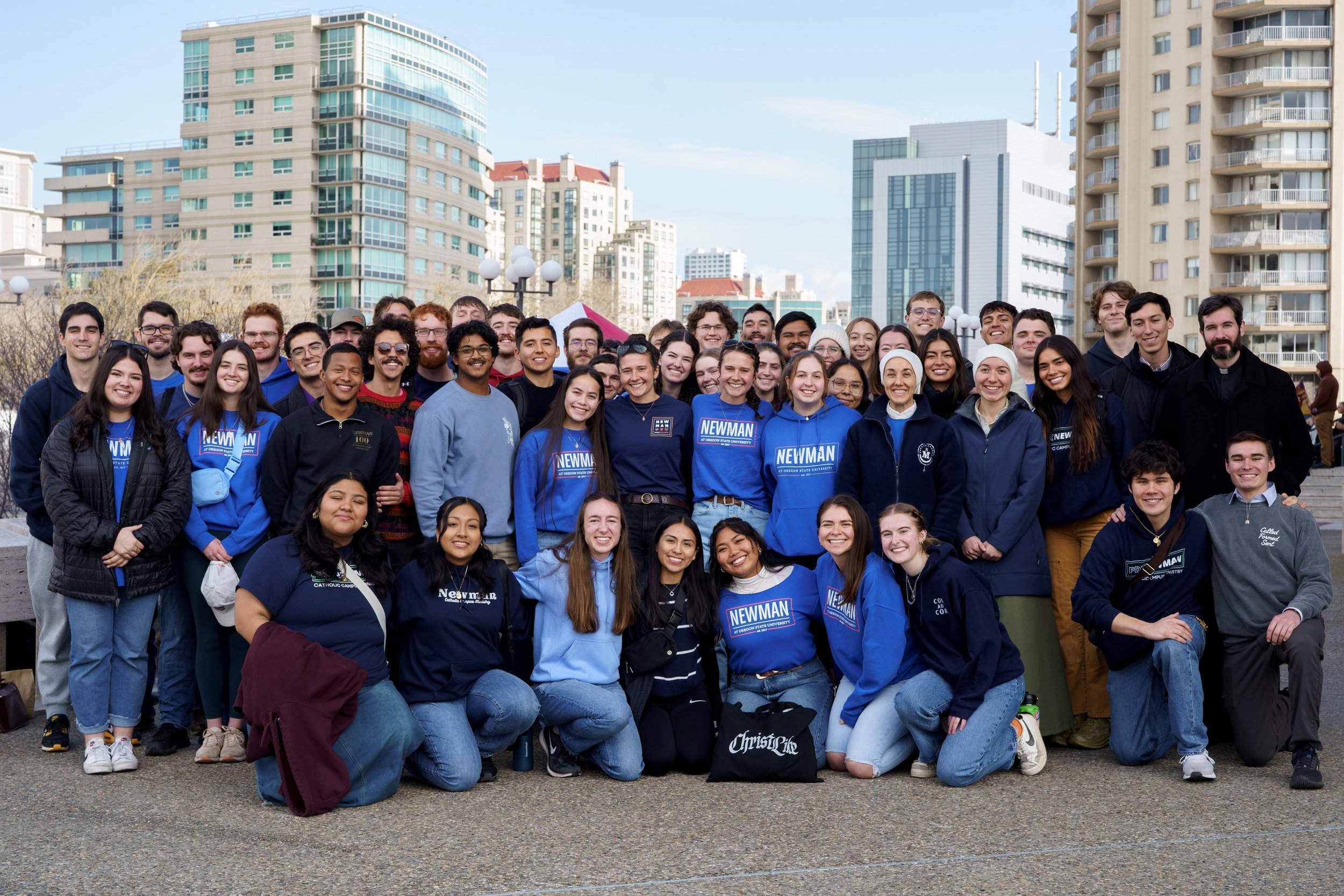 Group of diverse young adults and teenagers outdoors in an urban area with high-rise buildings in the background, smiling and posing for a photo.