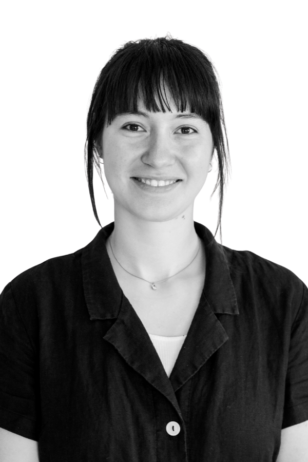 Black and white portrait of a woman with dark hair and bangs, smiling, wearing a dark button-up shirt and a small necklace, standing against a plain wall.