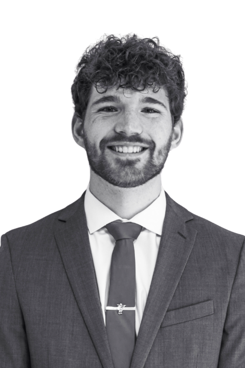 A black-and-white photo of a smiling man with curly hair, wearing a suit, dress shirt, and tie, standing against a plain background.