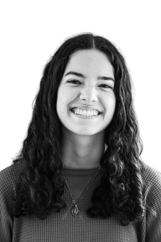 Black and white portrait of a smiling young woman with long dark hair, wearing earrings, a zipper vest, and a light-colored shirt, against a plain background.