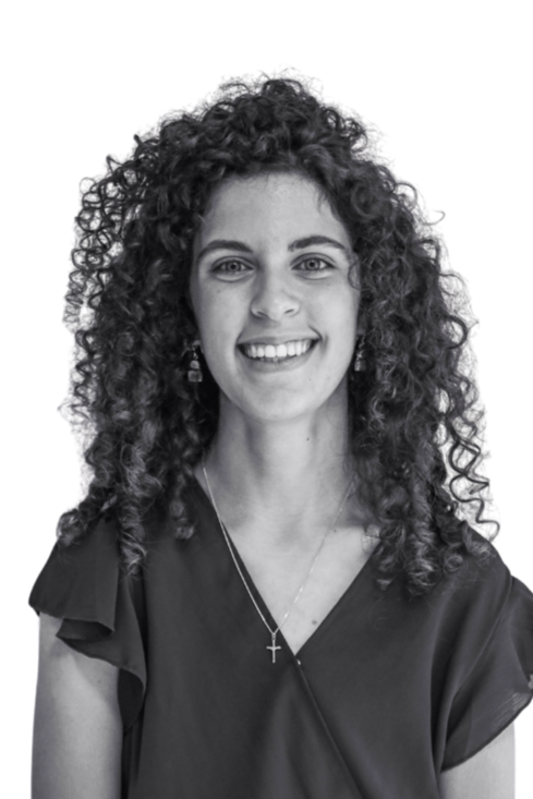 Black-and-white portrait of a smiling woman with curly hair, wearing earrings and a necklace with a cross pendant, standing against a plain wall.