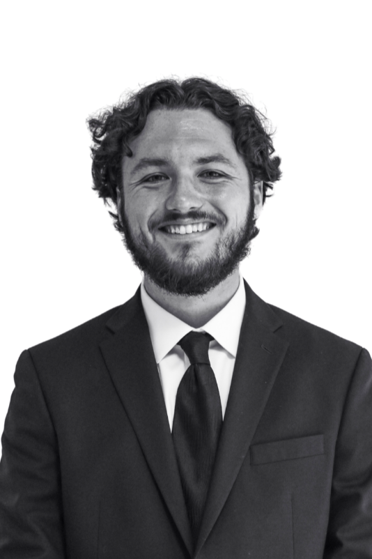 Black and white photo of a man with curly hair and a beard, smiling, dressed in a suit and tie, standing against a plain background.