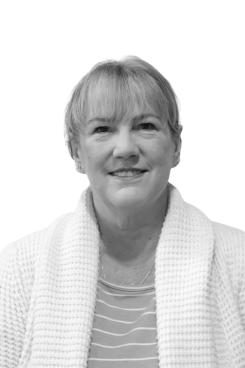 Black and white photo of a smiling middle-aged woman with short hair, wearing a striped shirt and a knitted sweater, standing against a plain background.