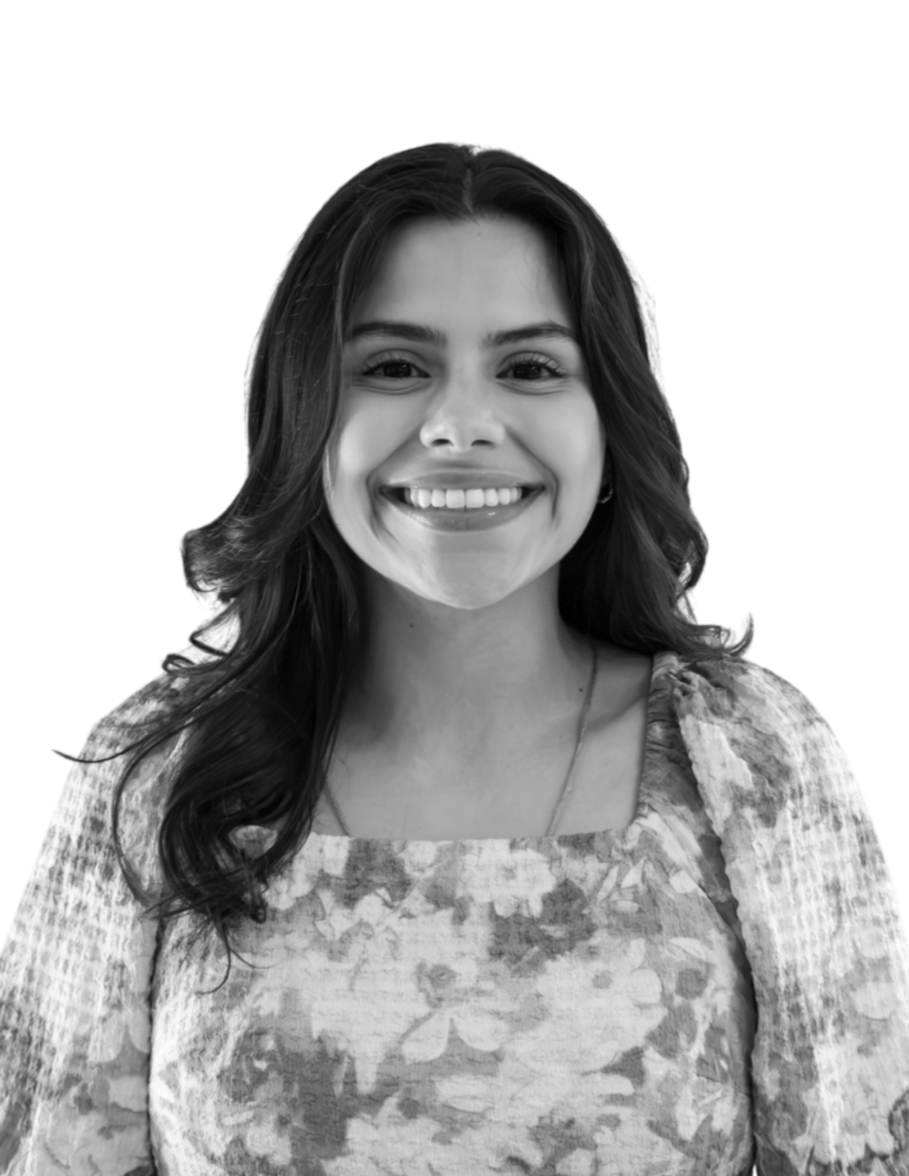Black-and-white portrait of a smiling woman with curly hair, wearing earrings and a necklace with a cross pendant, standing against a plain wall.