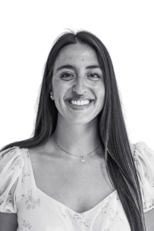 A woman with long dark hair, smiling, wearing a white floral top with puffy sleeves and a necklace, standing against a plain wall.