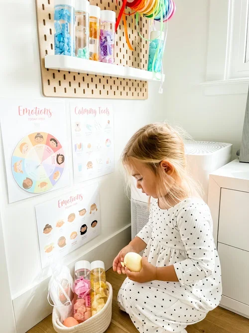 Photography of a toddler sitting in her calming corner with emotions posters and an emotions spinner wheel playing with sensory bottles and sensory stress balls