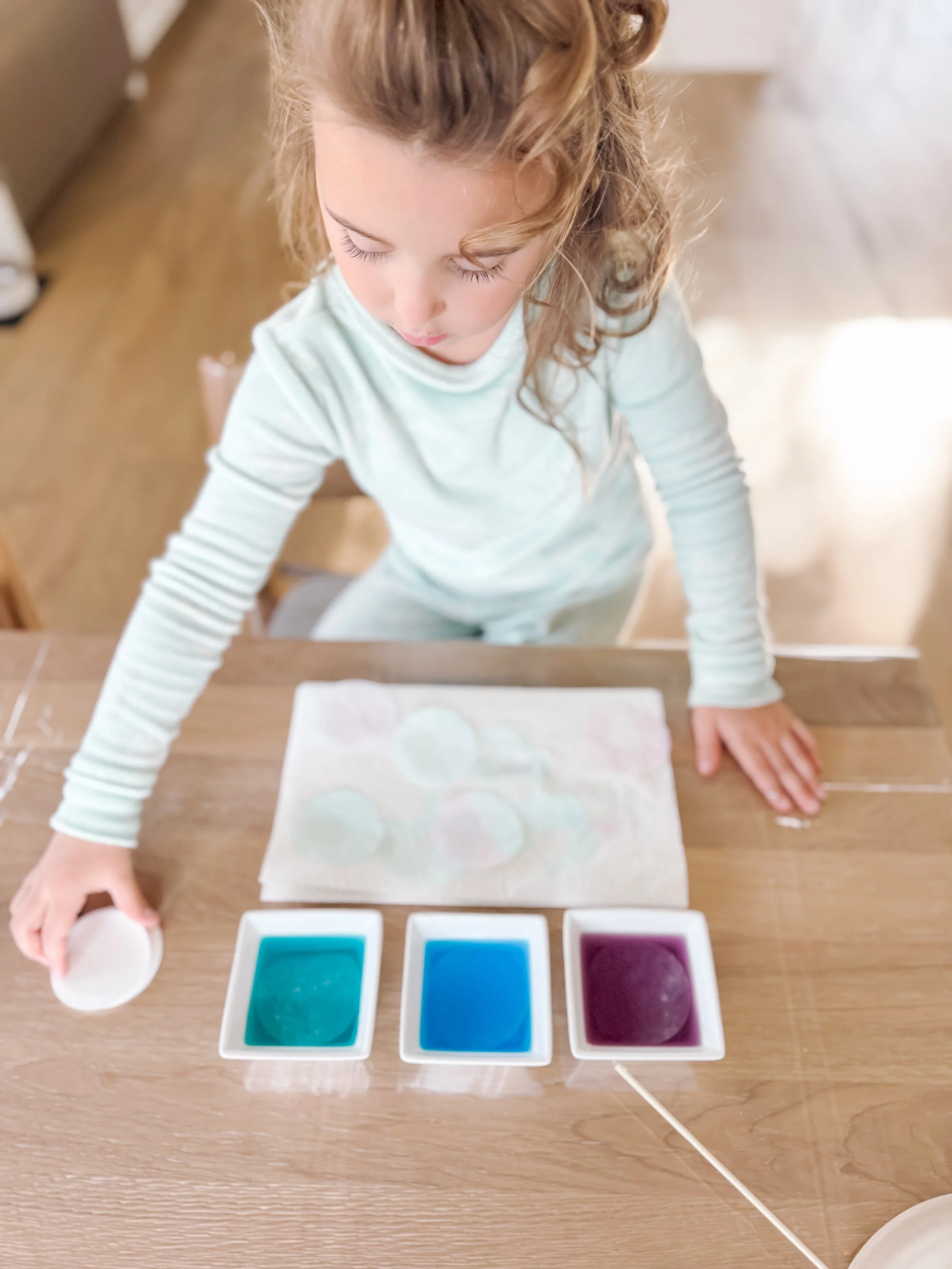  Materials for coffee filter paper flowers craft including white coffee filters, food coloring, water cups, craft sticks, and hot glue for toddler sensory play. 