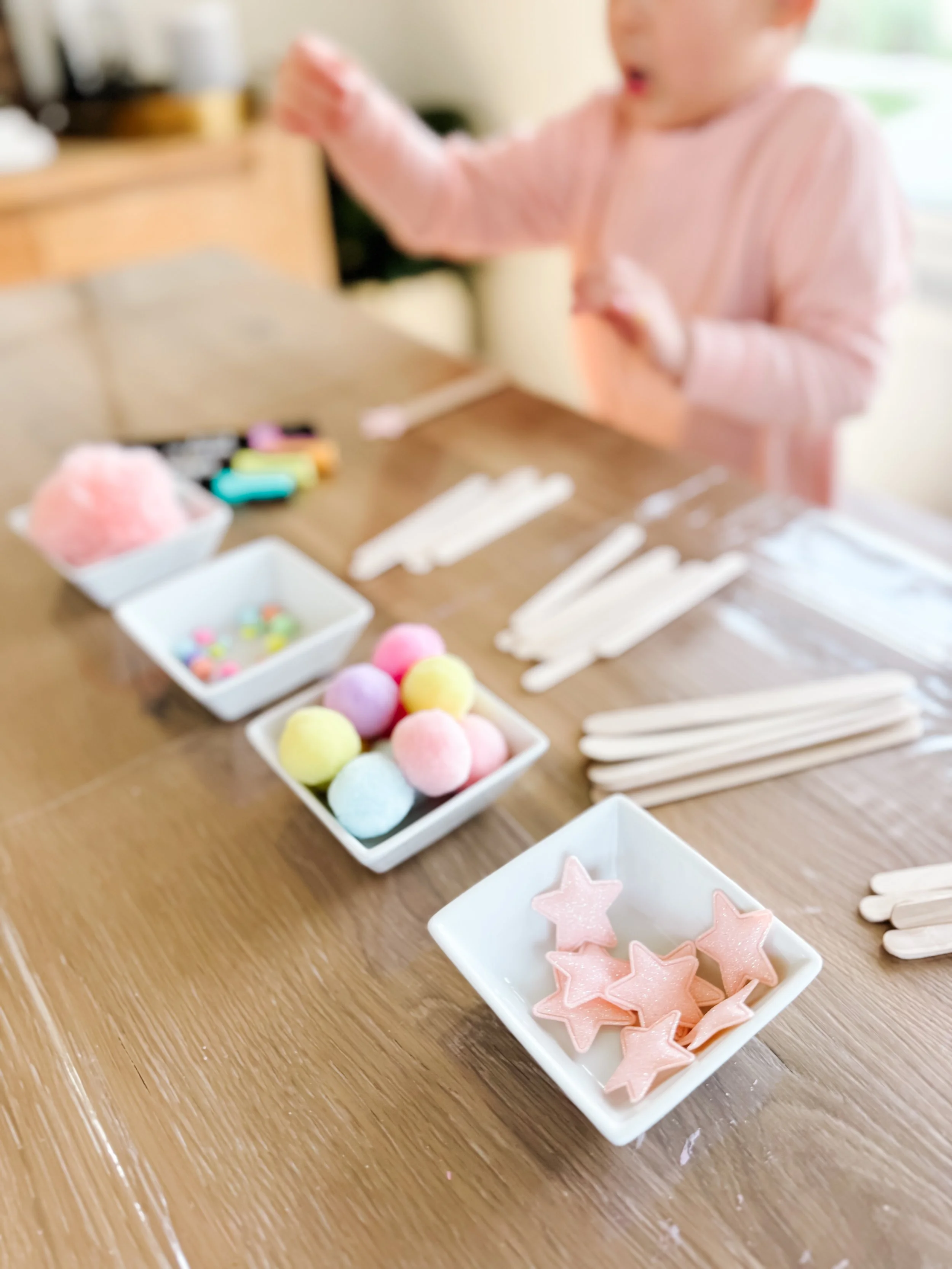 Pink glitter stars, pastel pom-poms, beads, craft sticks and yarn arranged for a children's weaving craft.