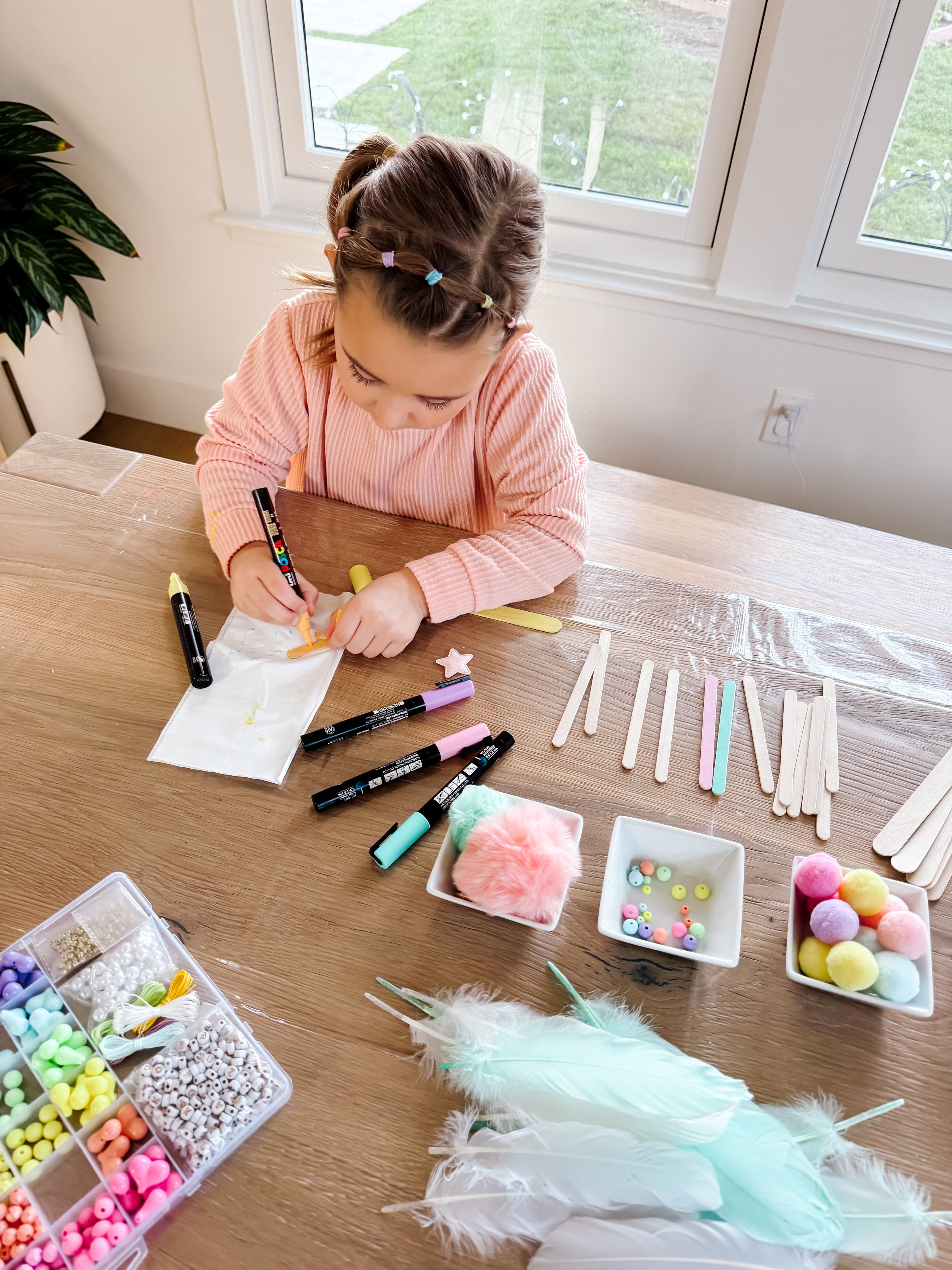 Child decorating wooden craft sticks with pastel paint markers for a dreamcatcher-inspired weaving craft.