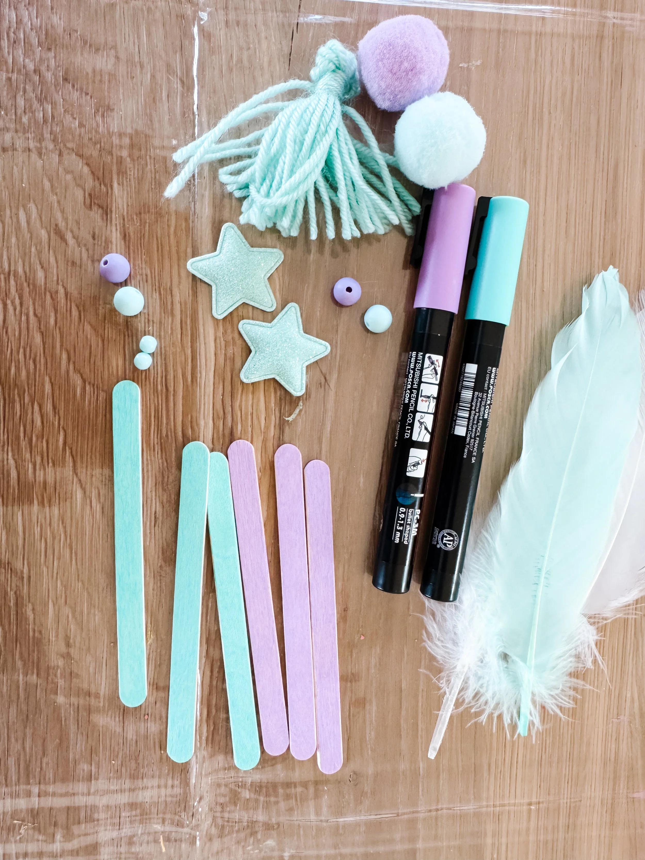 Mint and lavender craft sticks, yarn tassel, pom-poms, beads, stars and feathers for a cool-colored weaving craft.