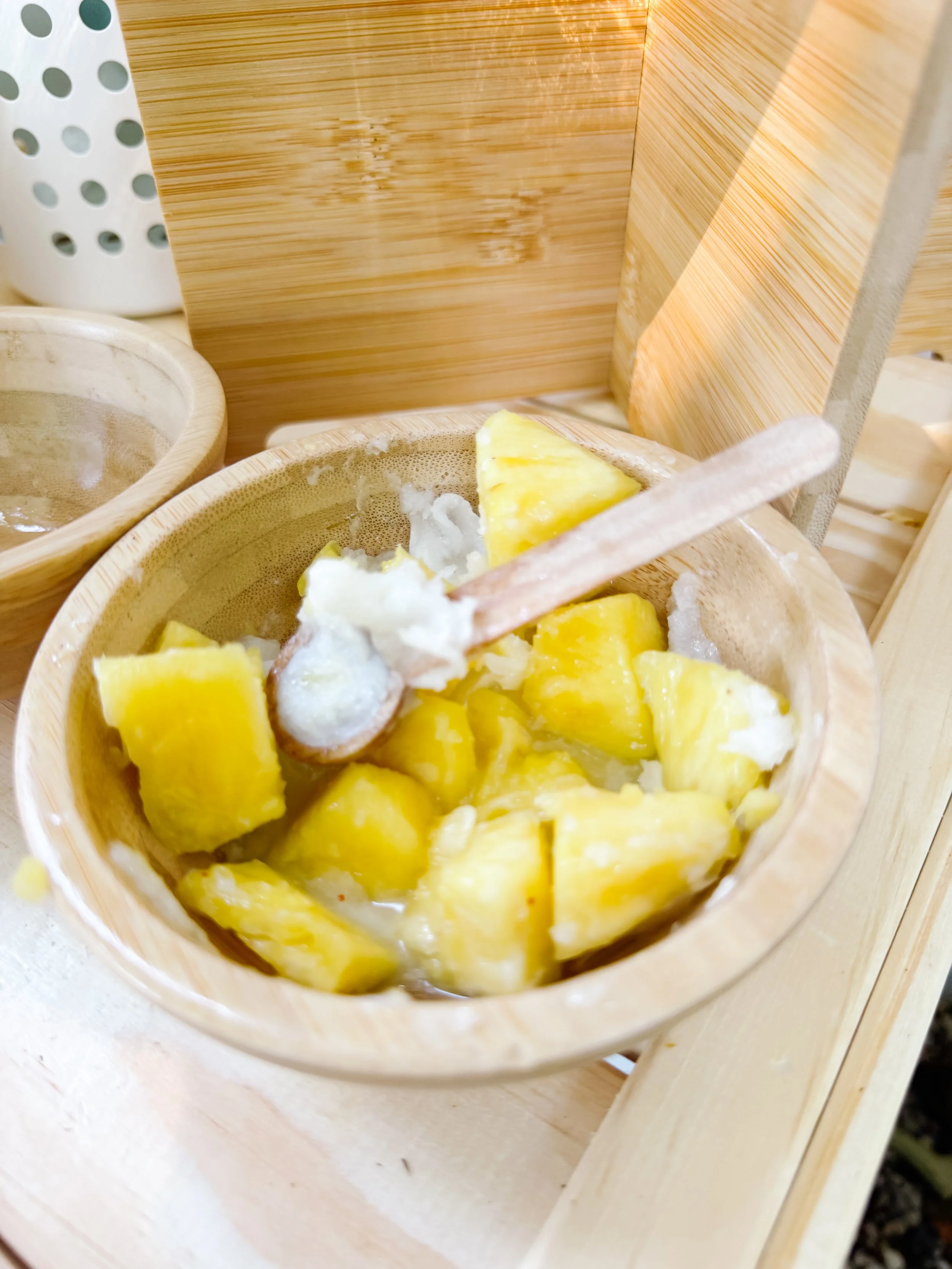 Child scooping pineapple and coconut into a wooden bowl for pretend smoothie play.
