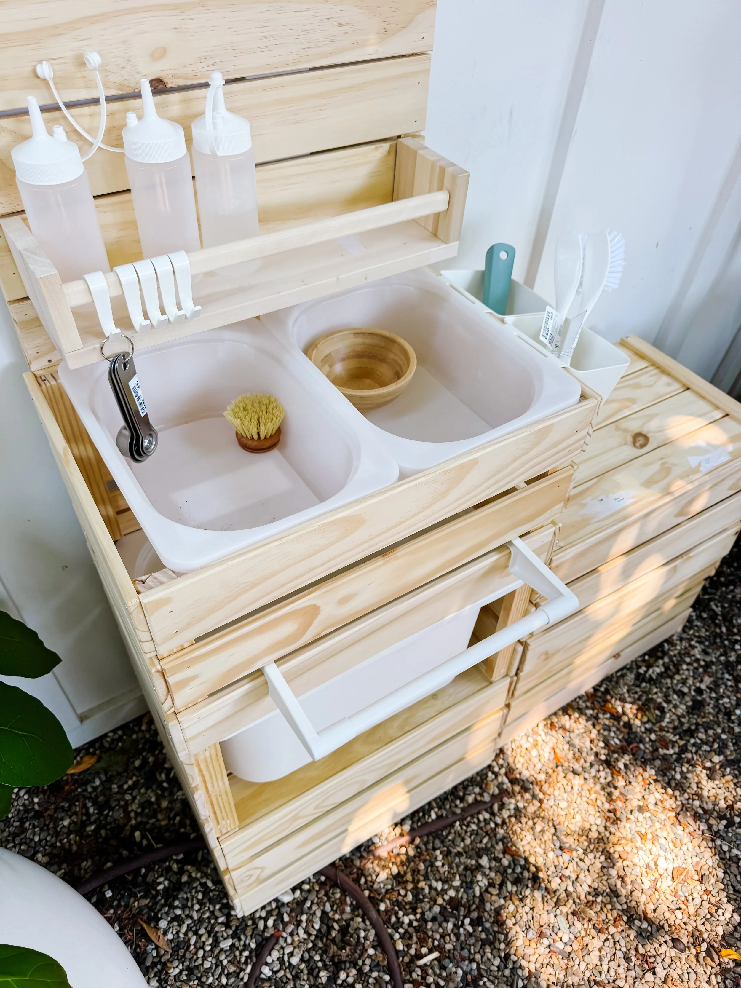 Outdoor mud kitchen setup with bowls, bottles, utensils, and sensory play supplies.
